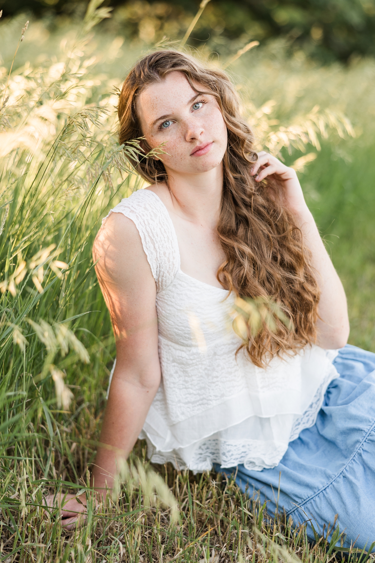 Mya, wearing a white lace top and long, flowy denim skirt, sits next to tall grass as the sun lights up the grass beautifully | CB Studio