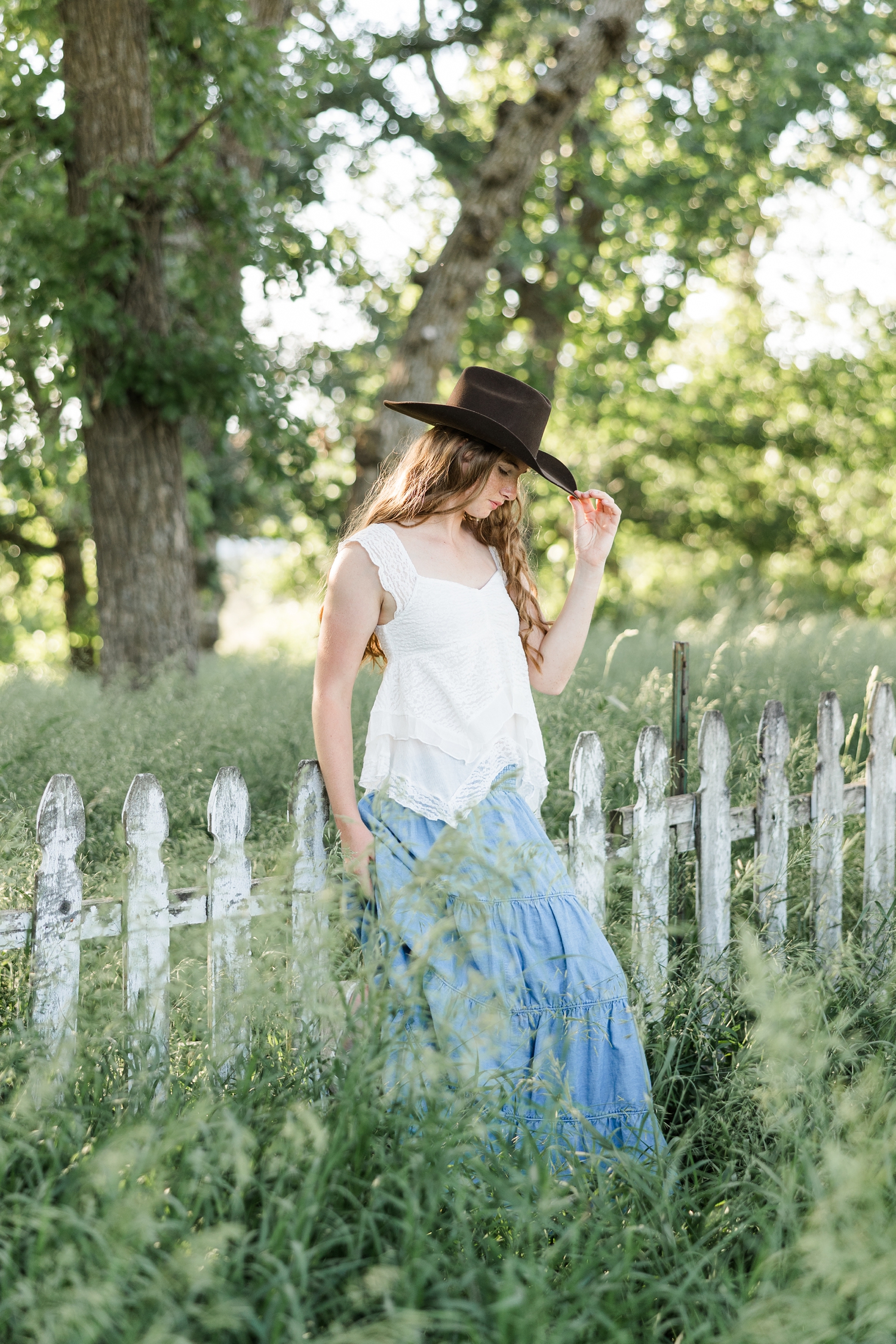 Mya, wearing a white lace top and long, flowy denim skirt and brown cowgirl hat, leans against rustic white picket fence in a grassy pasture as she tips her hat towards the camera | CB Studio