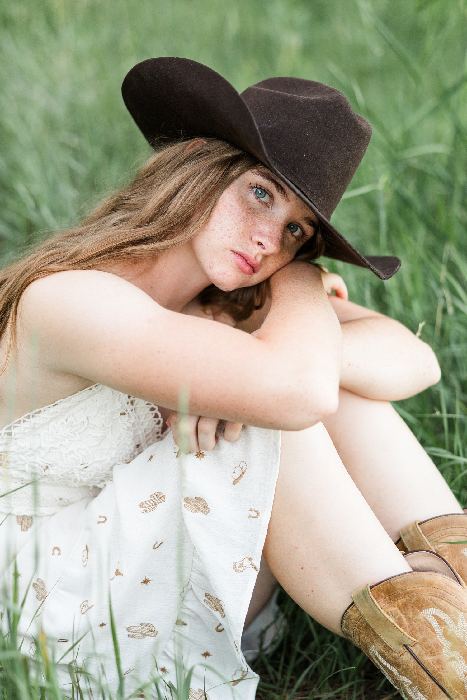 Mya, wearing a white dress with brown cowgirl print, brown cowgirl hat and brown boots, sits surrounded by tall grass with her knees up as she hugs them and rests her cheek on her arms | CB Studio