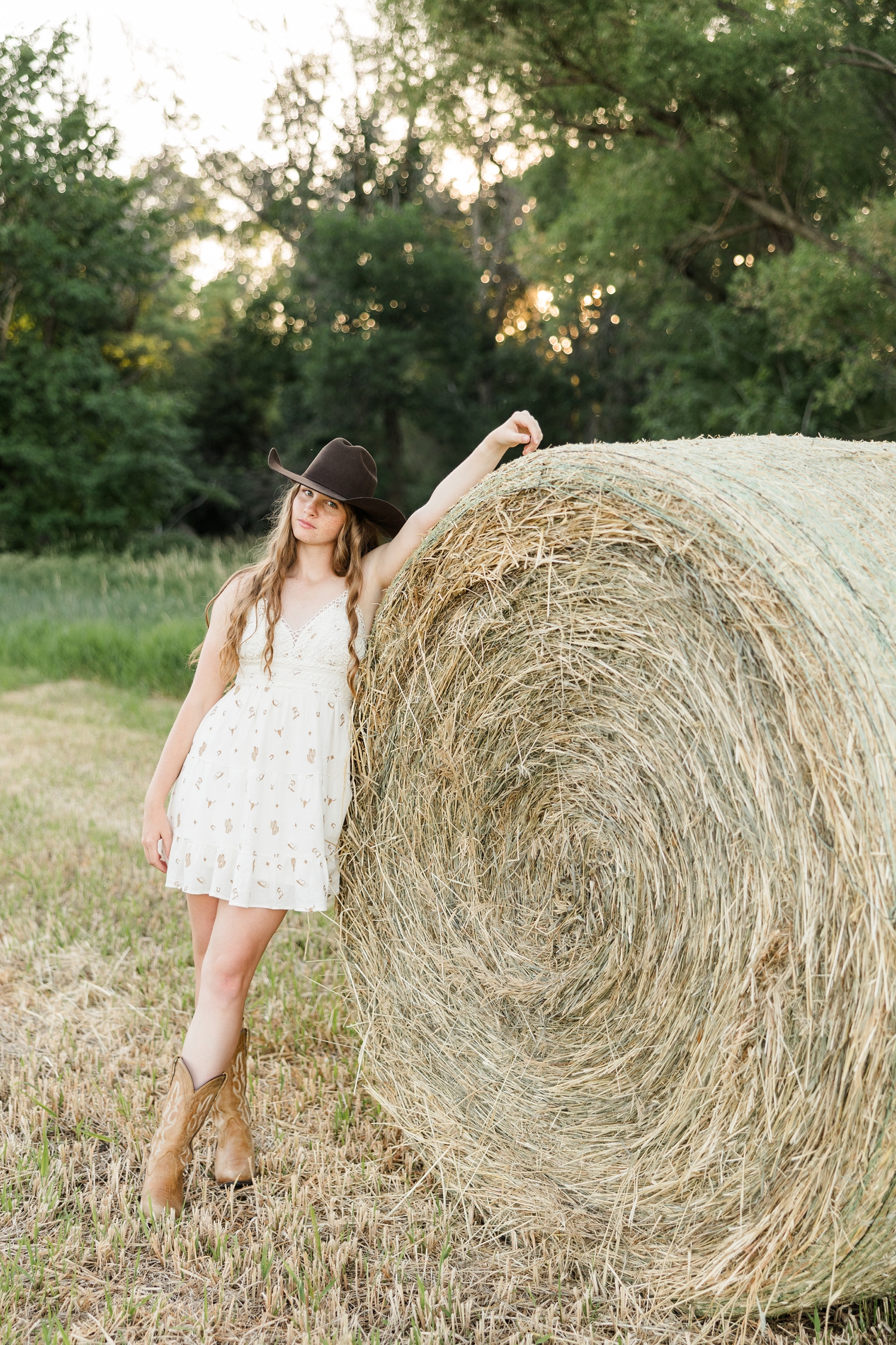 Mya, wearing a white dress with cowgirl boot print, brown cowgirl hat and brown boots, leans against a large round bale | CB Studio