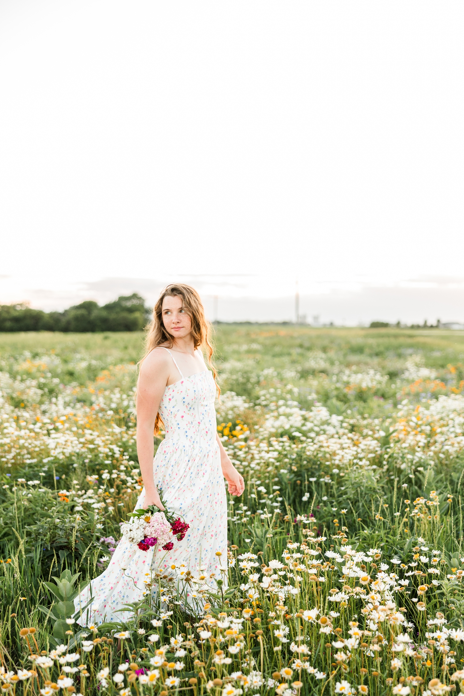 Mya, wearing a white, ditzy floral dress, walks slowly through a wildflower field at sunset while holding a small bouquet and looking back over her shoulder | CB Studio