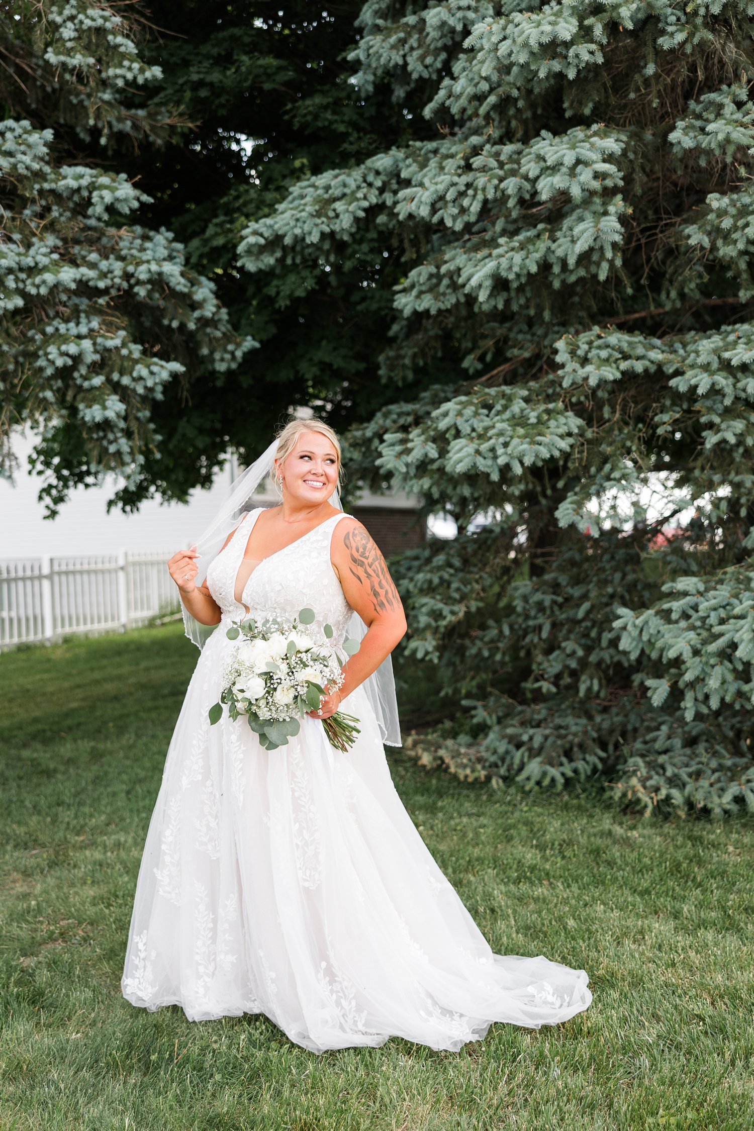 Teri stands beautifully among pine trees in her Essence of Australia wedding gown as she looks off to the right and gentle runs her fingers down her veil | CB Studio
