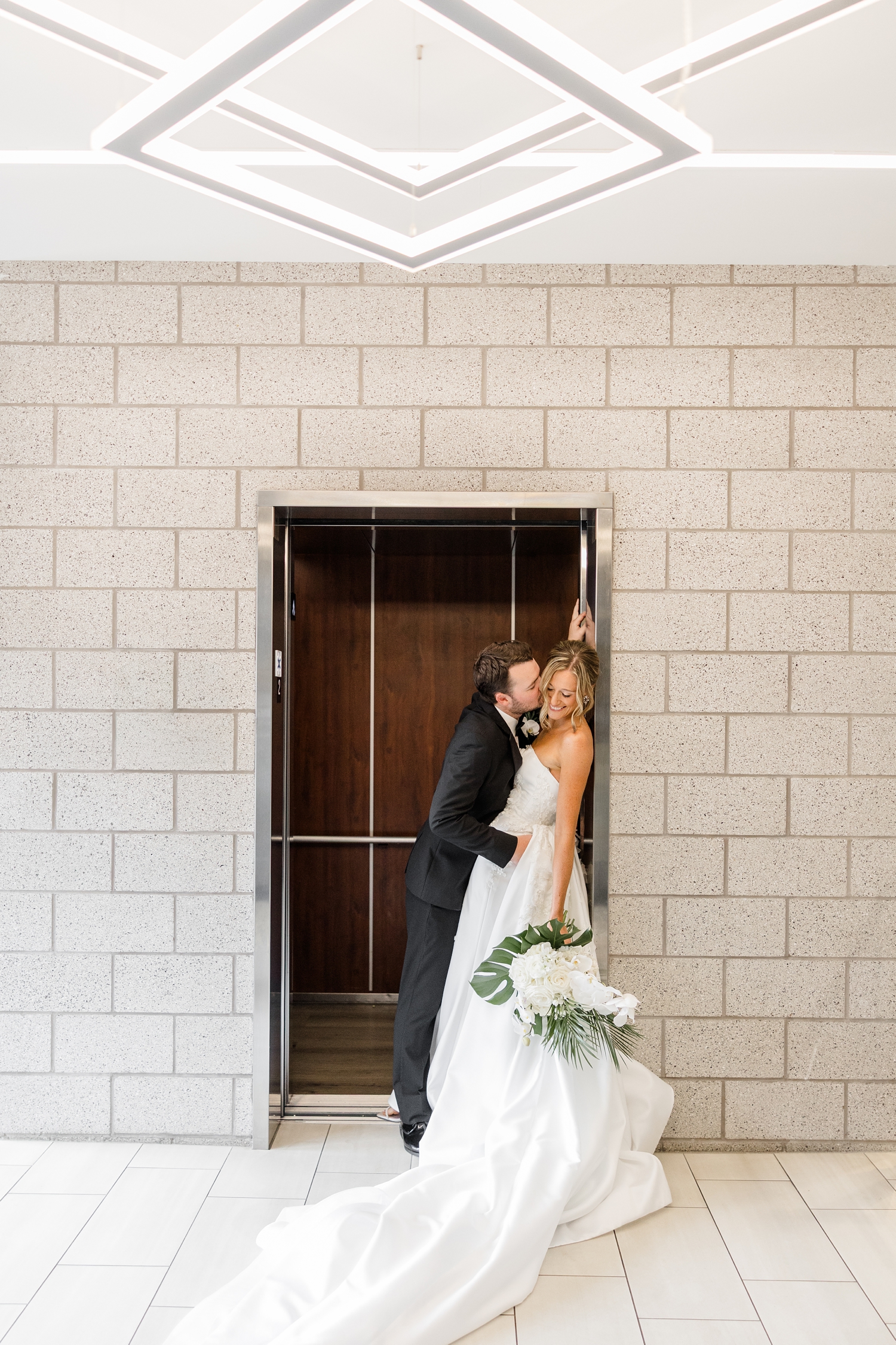 Cassidy leans against one side of the door opening of the elevator while Grant leans in to kiss her cheek at Toast in Ankeny, IA | CB studio