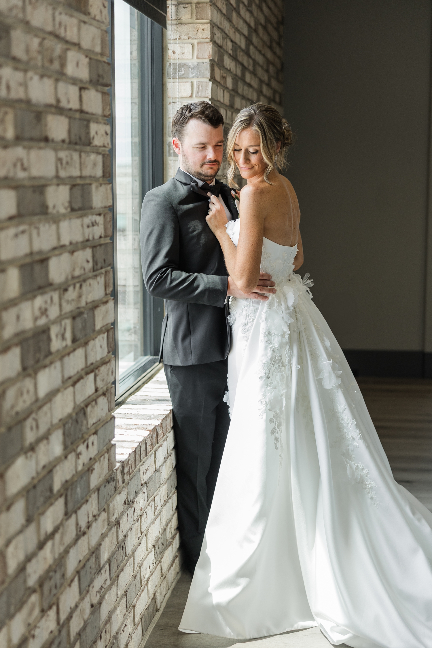 Grant pulls his bride, Cassidy, in close and they both look down while leaning against a brick wall and window at Toast in Ankeny, IA | CB Studio