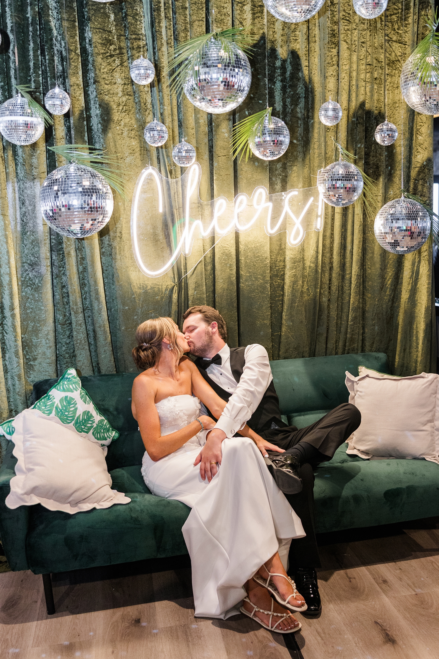 Cassidy shares a kiss with her new husband, Grant, as they sit on a green velvet couch under a green velvet backdrop decorated with disco balls and a neon "cheers" sign. | CB Studio