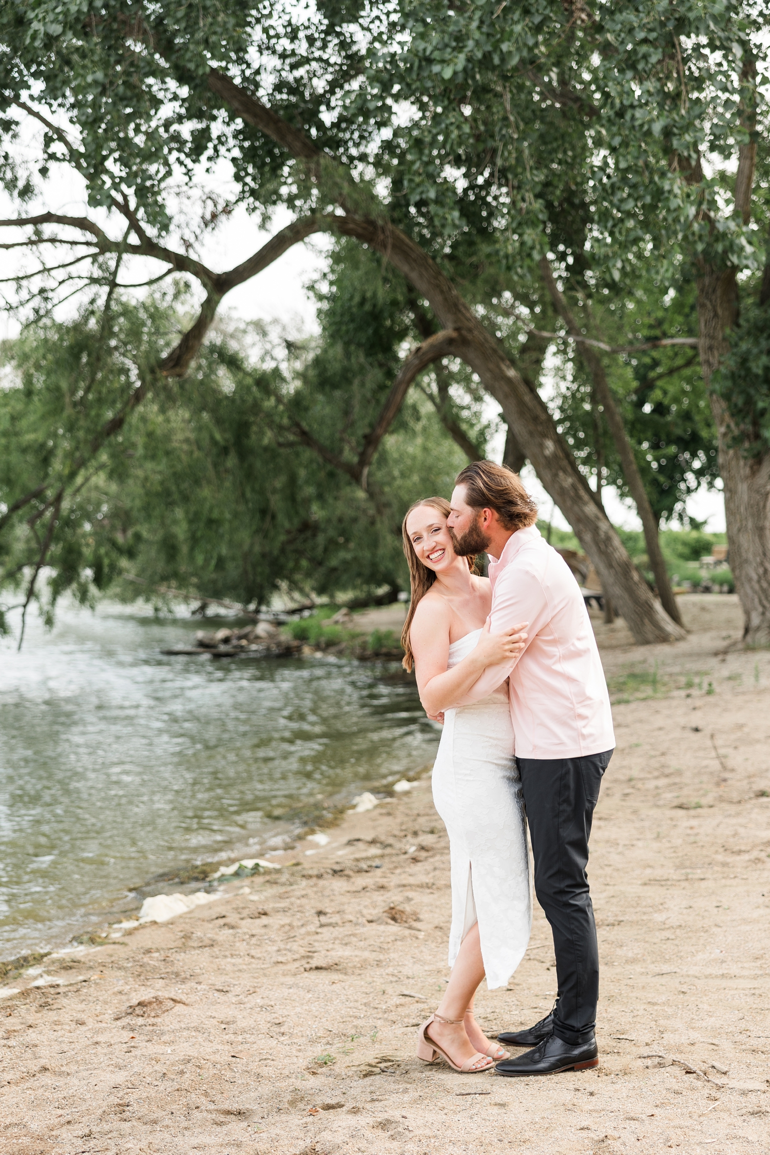 Zach embraces Chloe and kisses her cheek as she laughs on the beach of Lake Cornelia | CB Studio