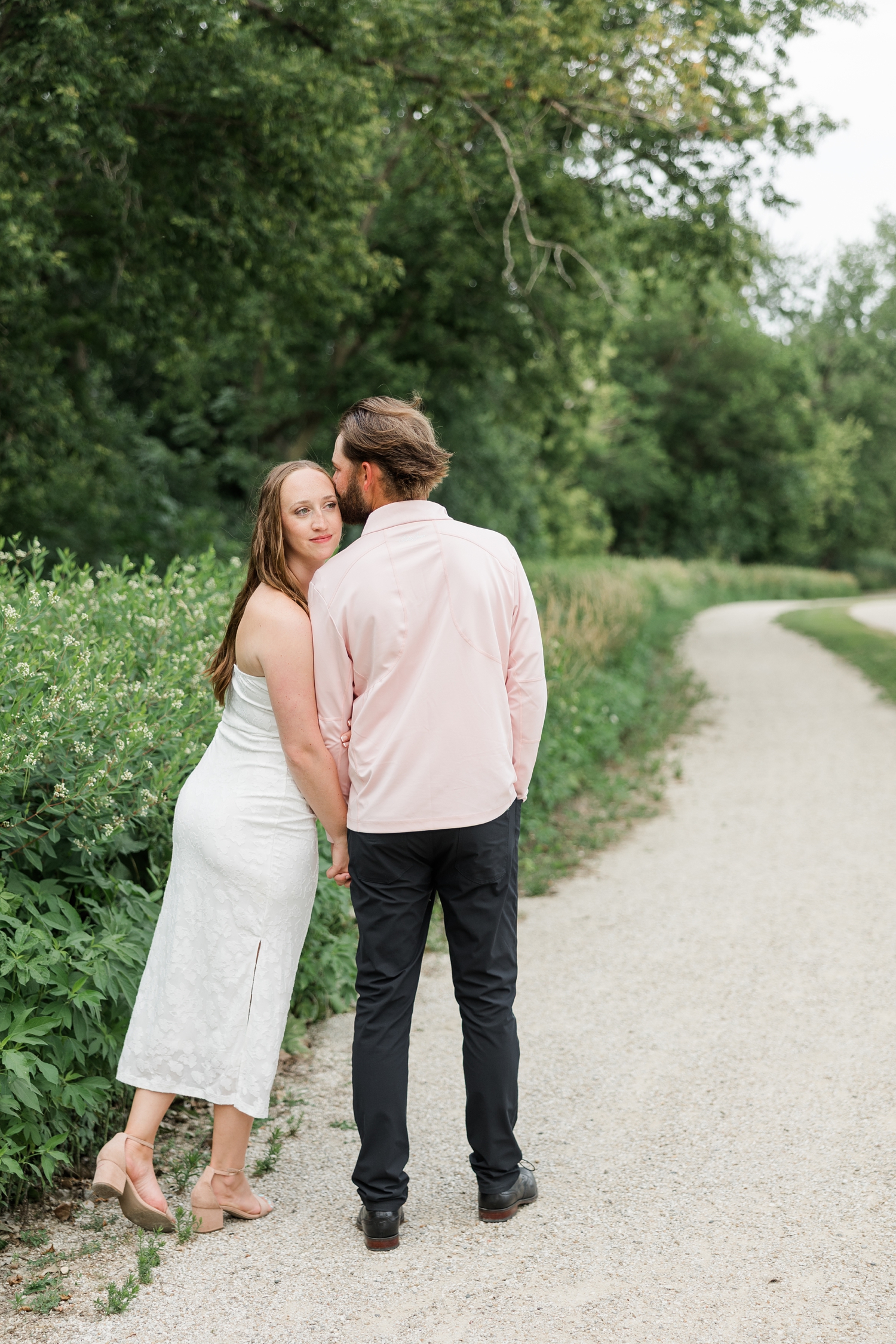 Chloe looks behind Zach's shoulder has he kisses her forehead as they walk down a path near Lake Cornelia | CB Studio
