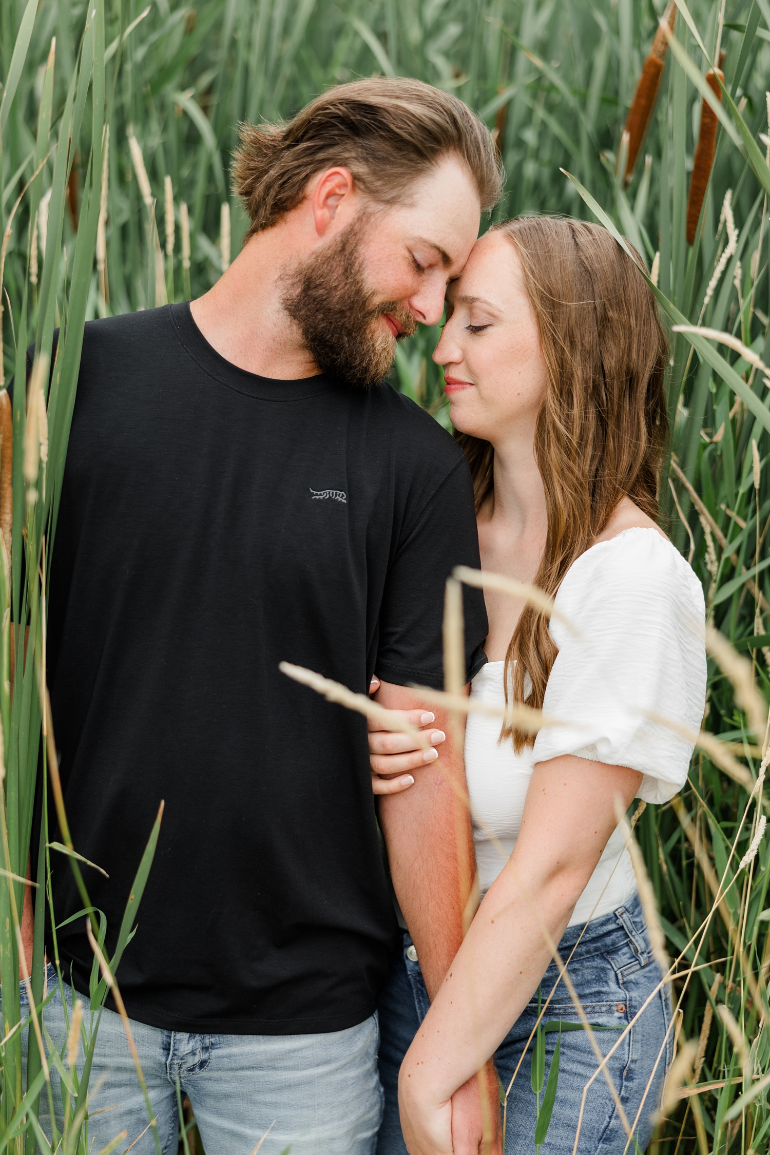 In a patch of tall cat tail grass, Chloe stands slightly behind Zach as they hold hands and rest their foreheads together | CB Studio