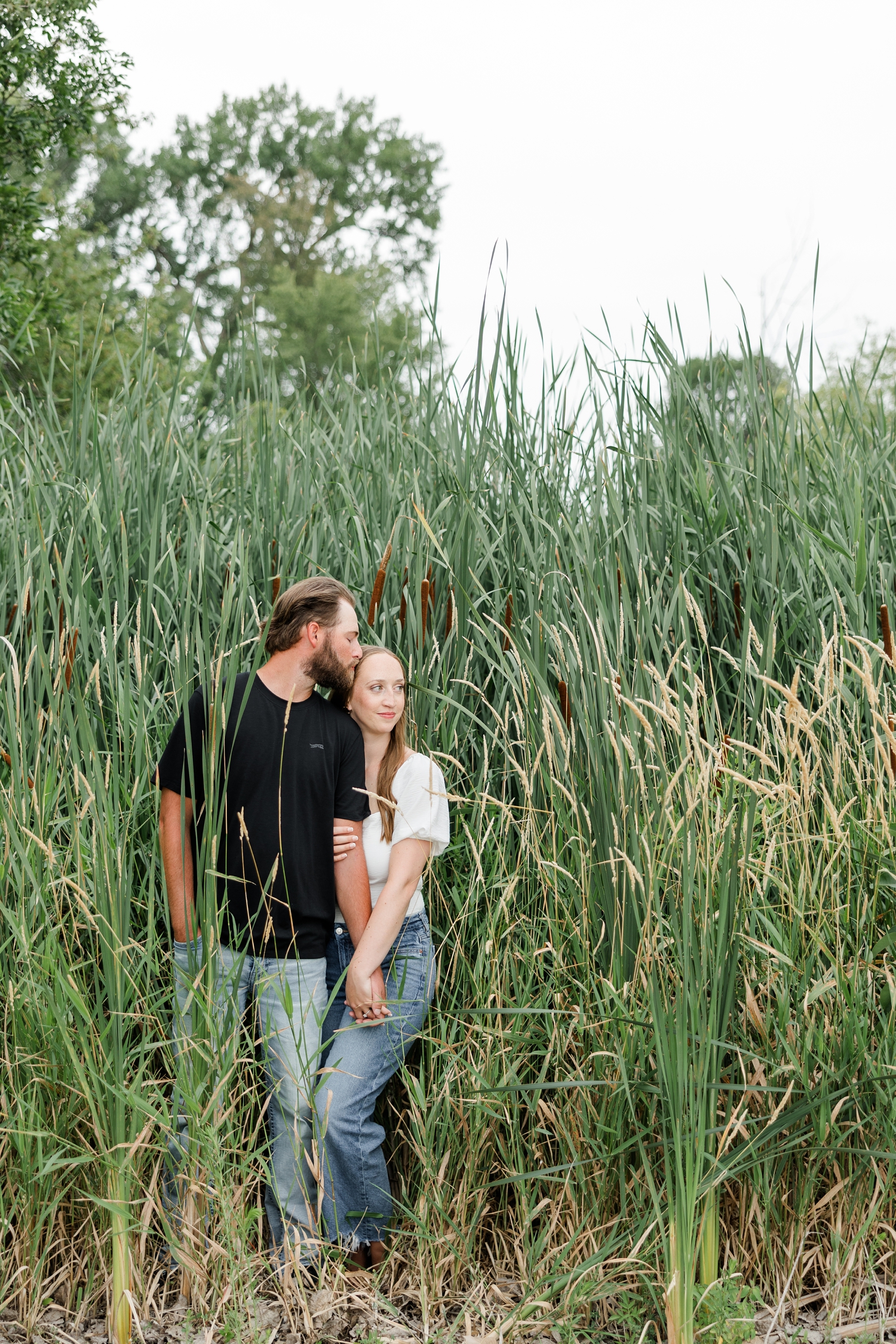 In a patch of tall cat tail grass, Chloe stands slightly behind Zach as they hold hands and Zach kisses her forehead | CB Studio