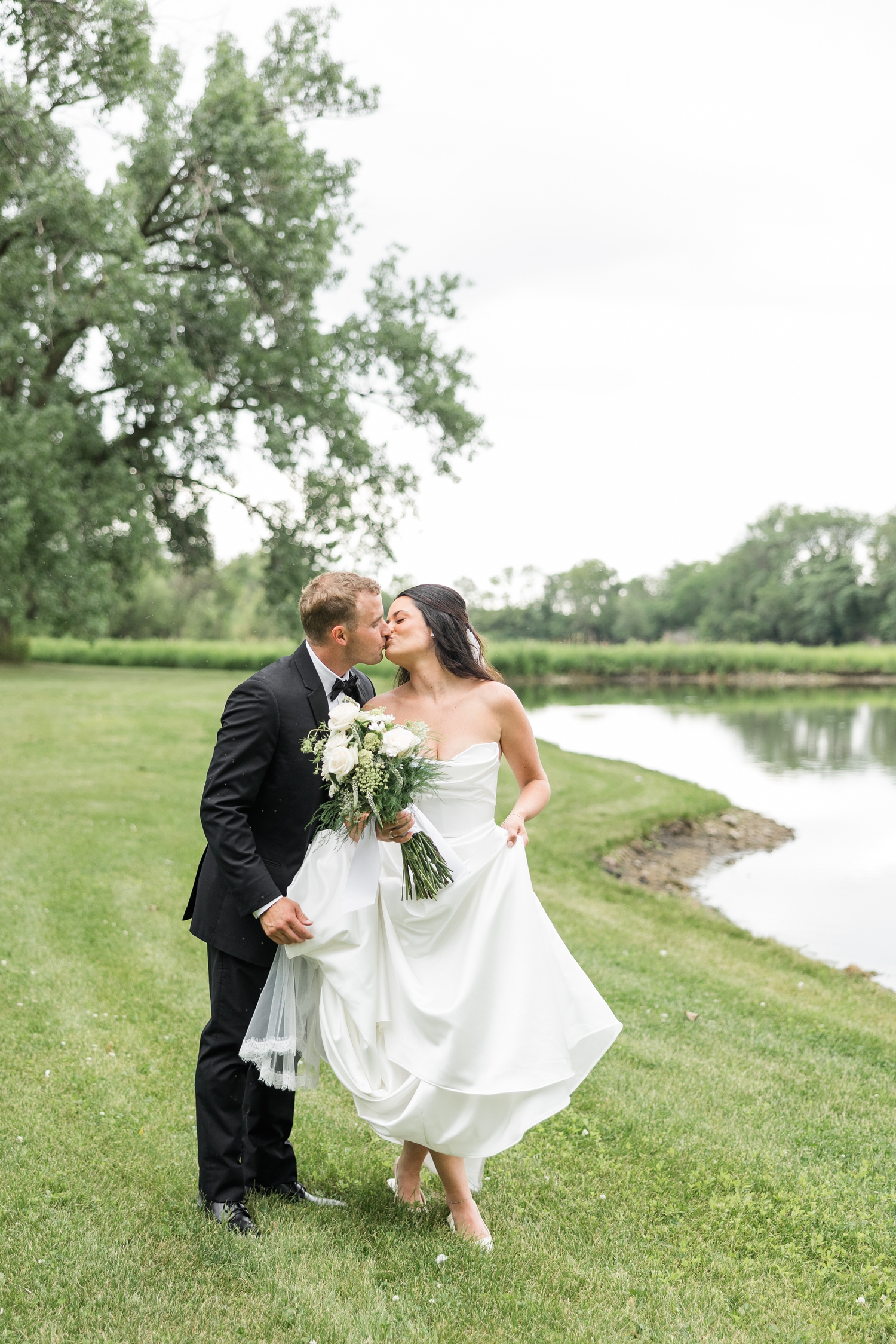 Bride and groom, Emily and Derrick, pause to share a kiss as they walk along a pond while Derrick helps hold Emily's train of her wedding gown. | CB Studio