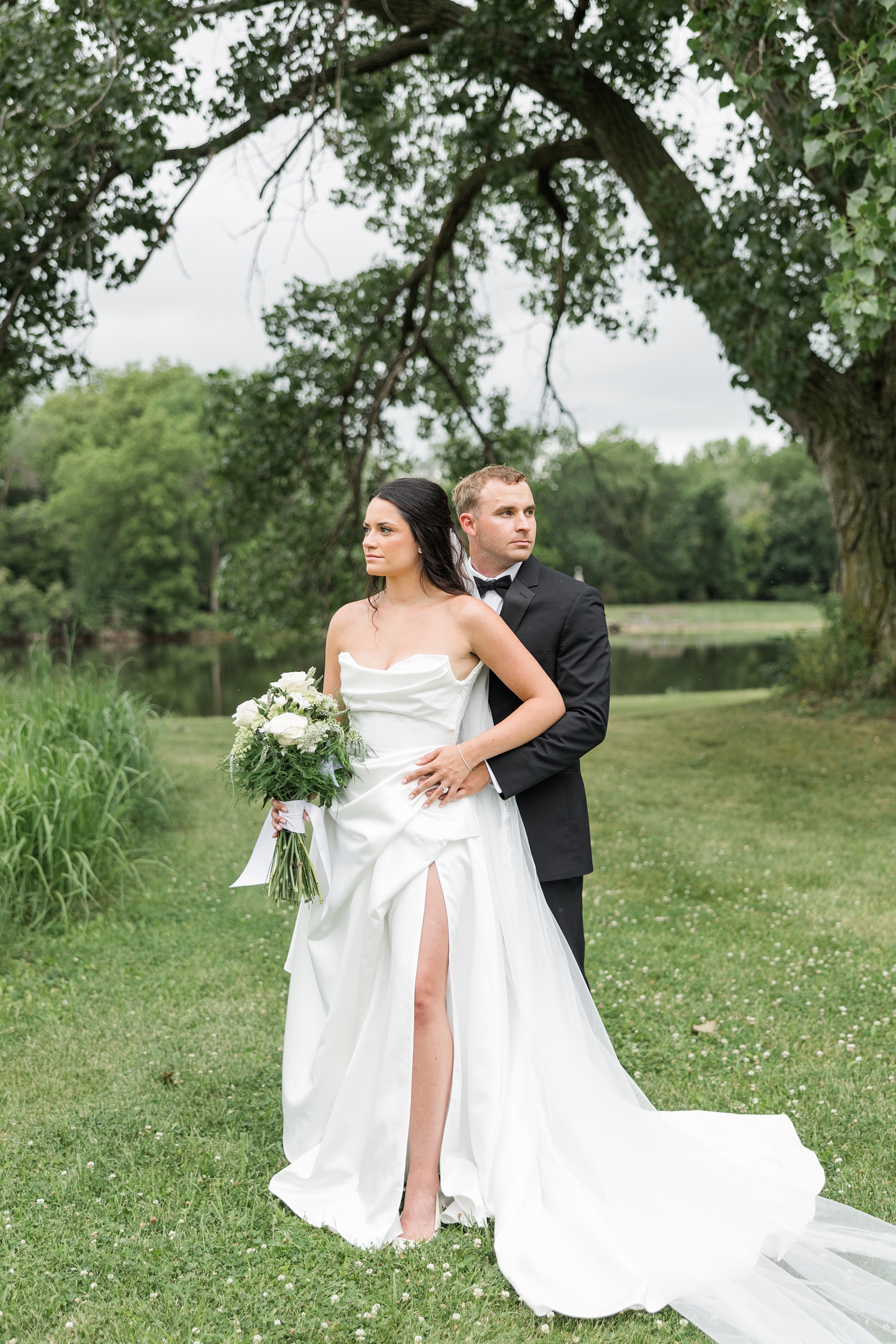 Bride and groom, Emily and Derrick, stand in a beautiful pond landscape as Emily looks to the left and Derrick to the right | CB Studio