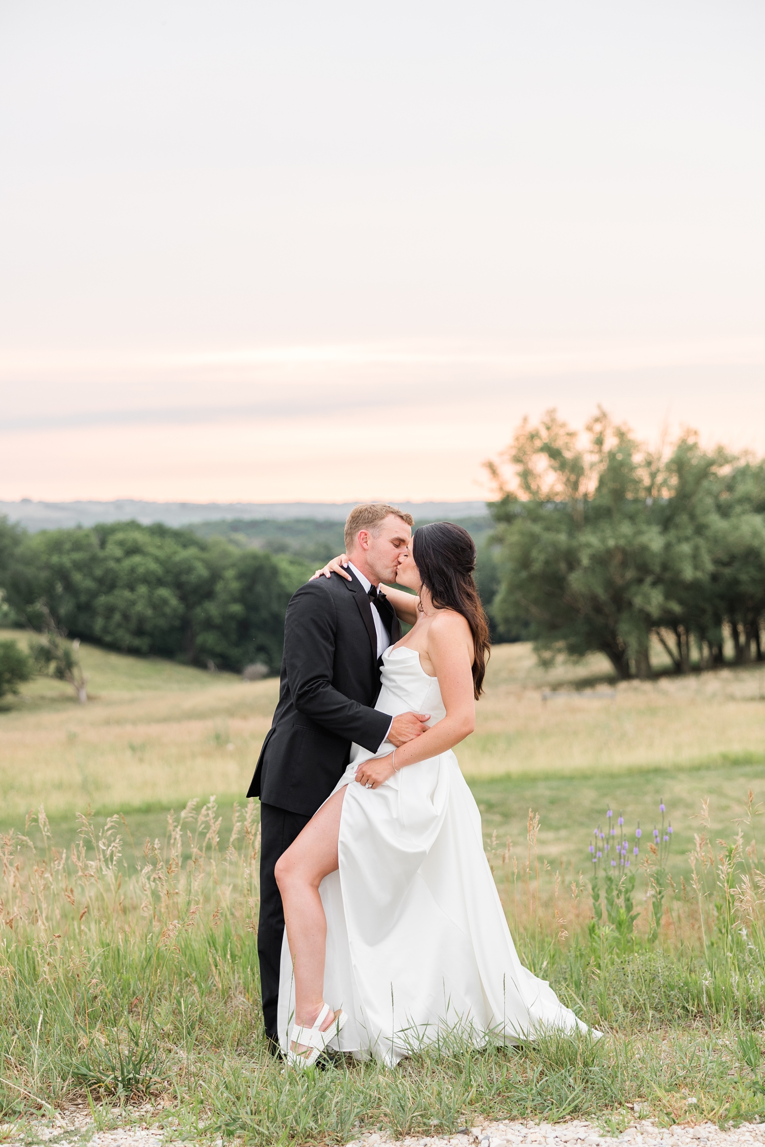 Derrick leans his bride, Emily, back for a kiss with a hilly pasture and beautiful sunset in the background at The Stables at Copper Ridge | CB Studio