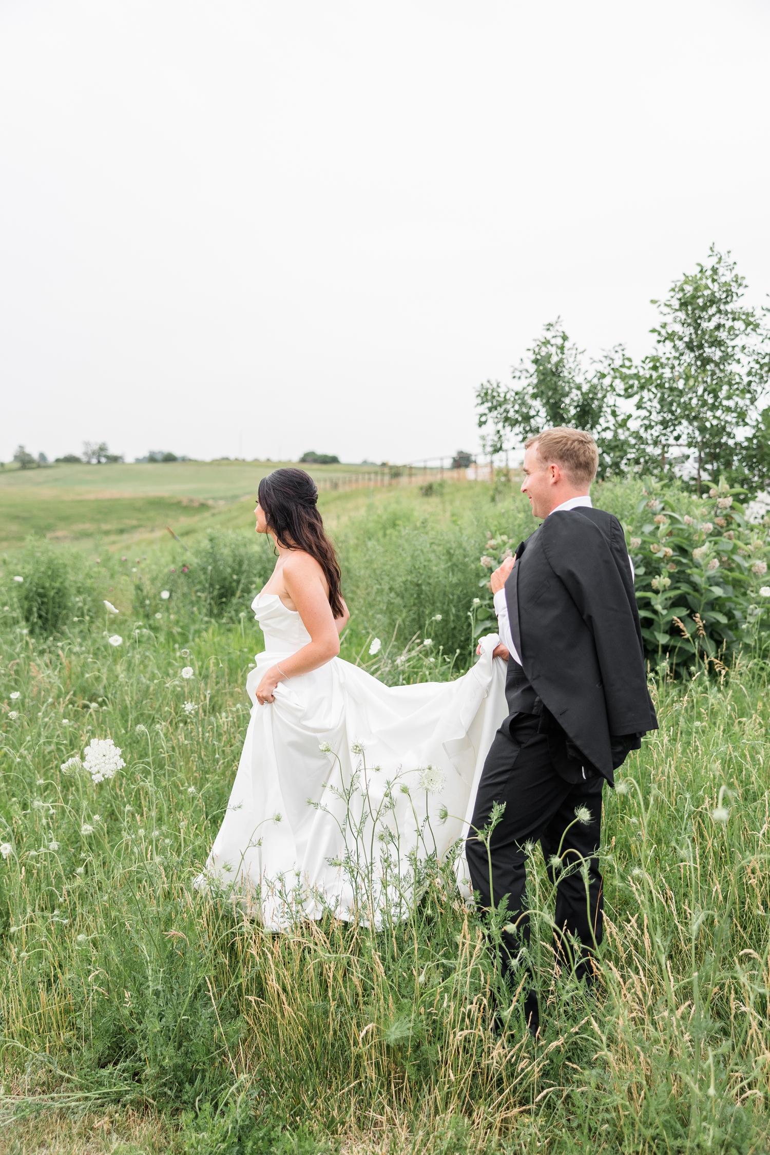 Emily walks through a hilly, grassy, flower field while her groom, Derrick, follows behind holding the train of her wedding gown | CB Studio