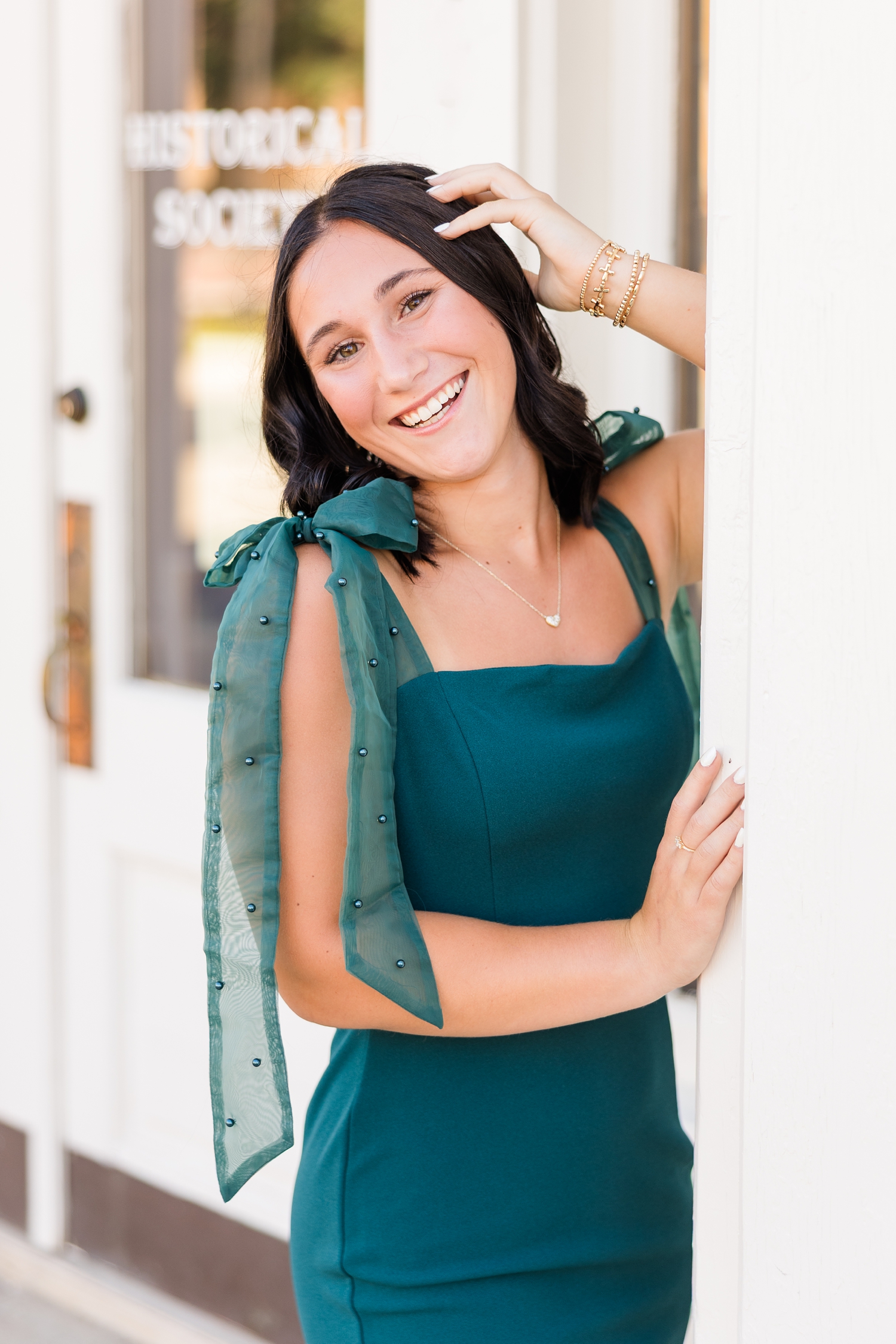 Ivy, wearing an emerald body-con dress with bow tie straps adorned with peals, leans against a white historical building in downtown Algona, IA | CB Studio