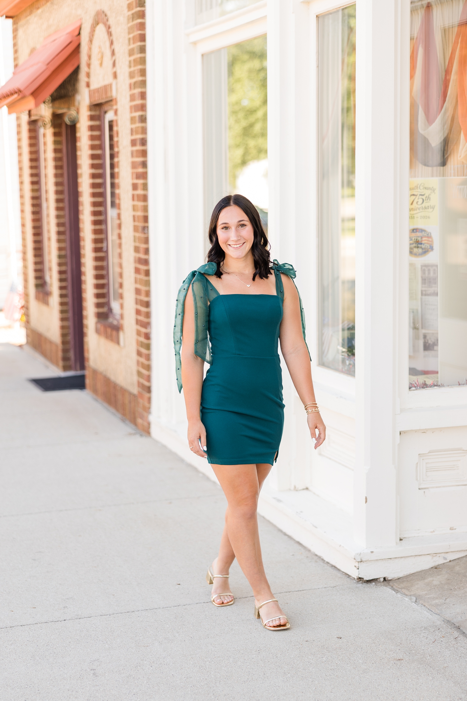 Ivy, wearing an emerald body-con dress with bow tie straps adorned with peals, walks down a sidewalk in downtown Algona, IA | CB Studio