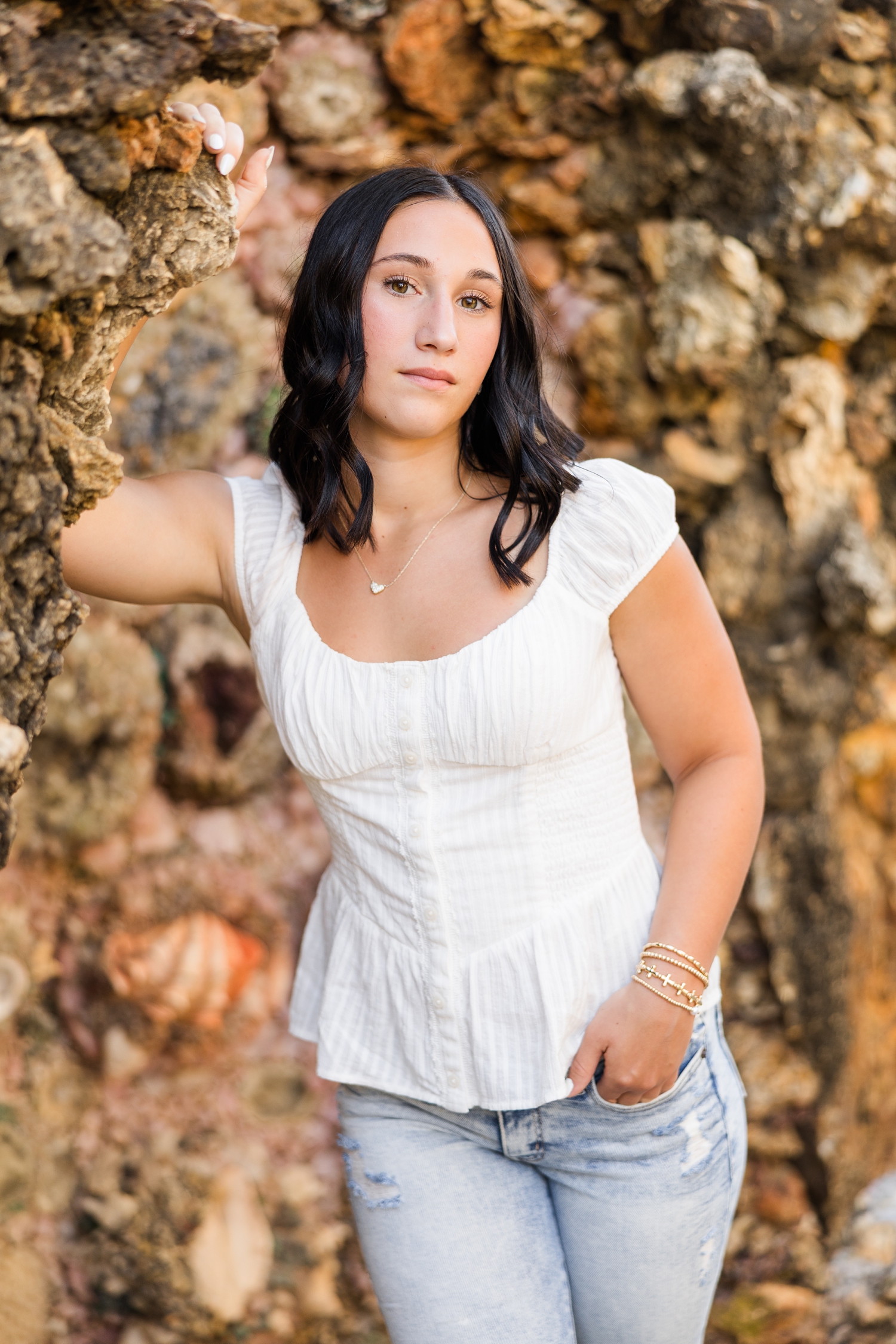 Ivy, wearing a white top and jeans, leans against a rock wall at The Grotto of Redemption | CB Studio