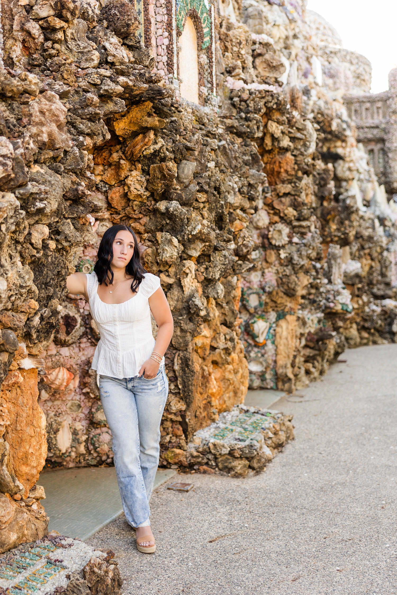 Ivy, wearing a white top and jeans, leans against a rock wall at The Grotto of Redemption while looking off to the right | CB Studio