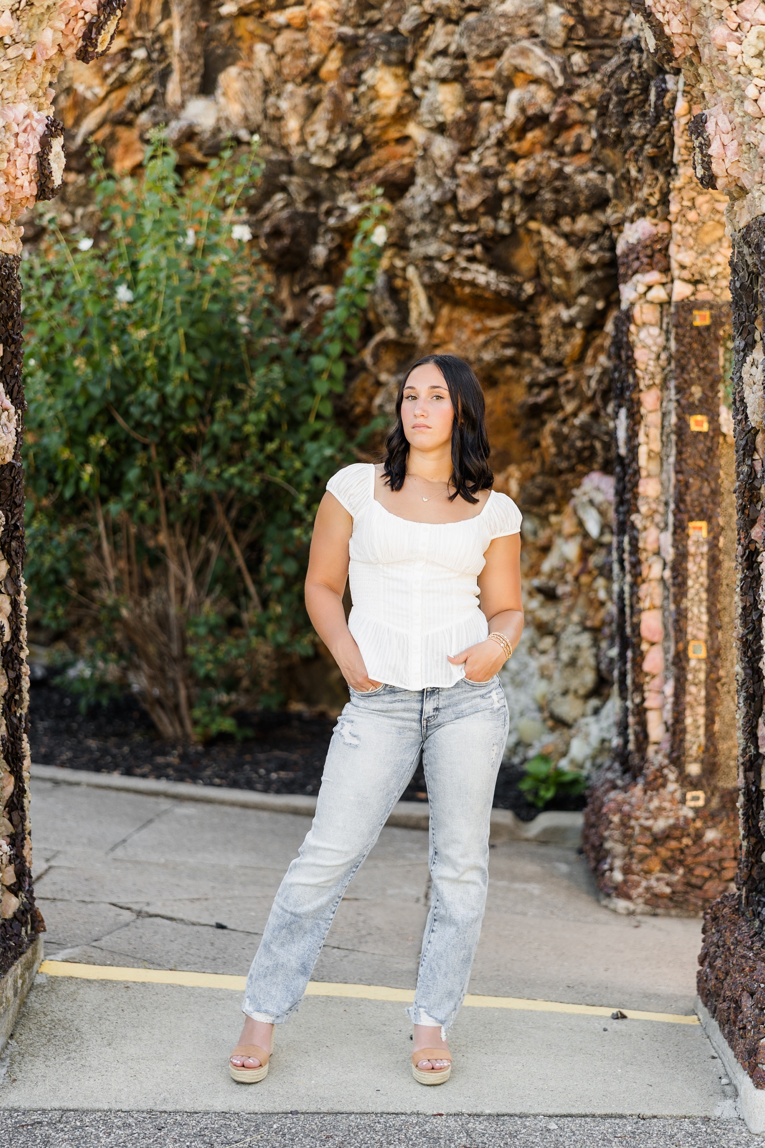 Ivy, wearing a white top and jeans, stands with her hands in her pockets in the middle of a rock archway at The Grotto of Redemption | CB Studio
