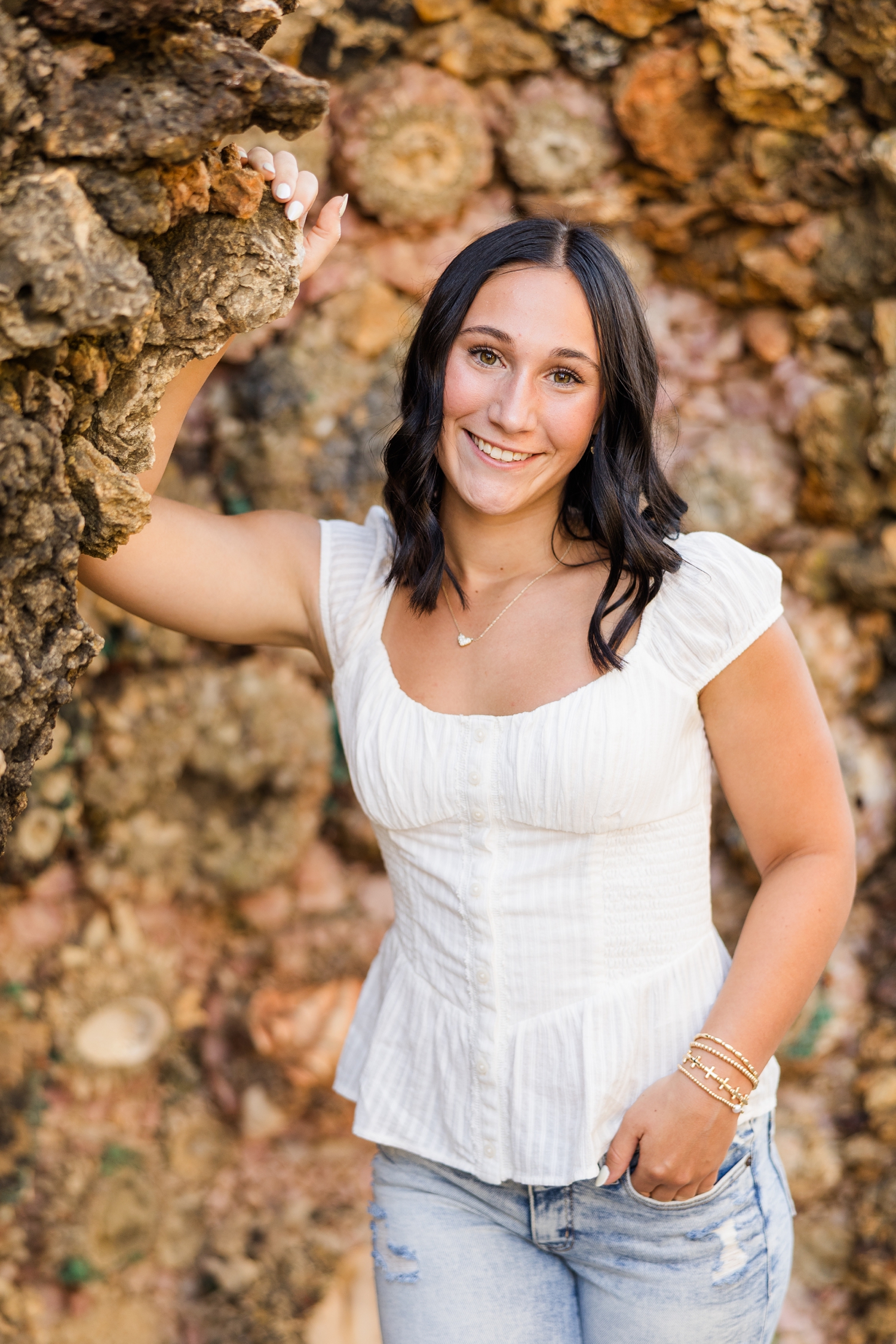 Ivy, wearing a white top and jeans, leans against a rock wall at The Grotto of Redemption | CB Studio