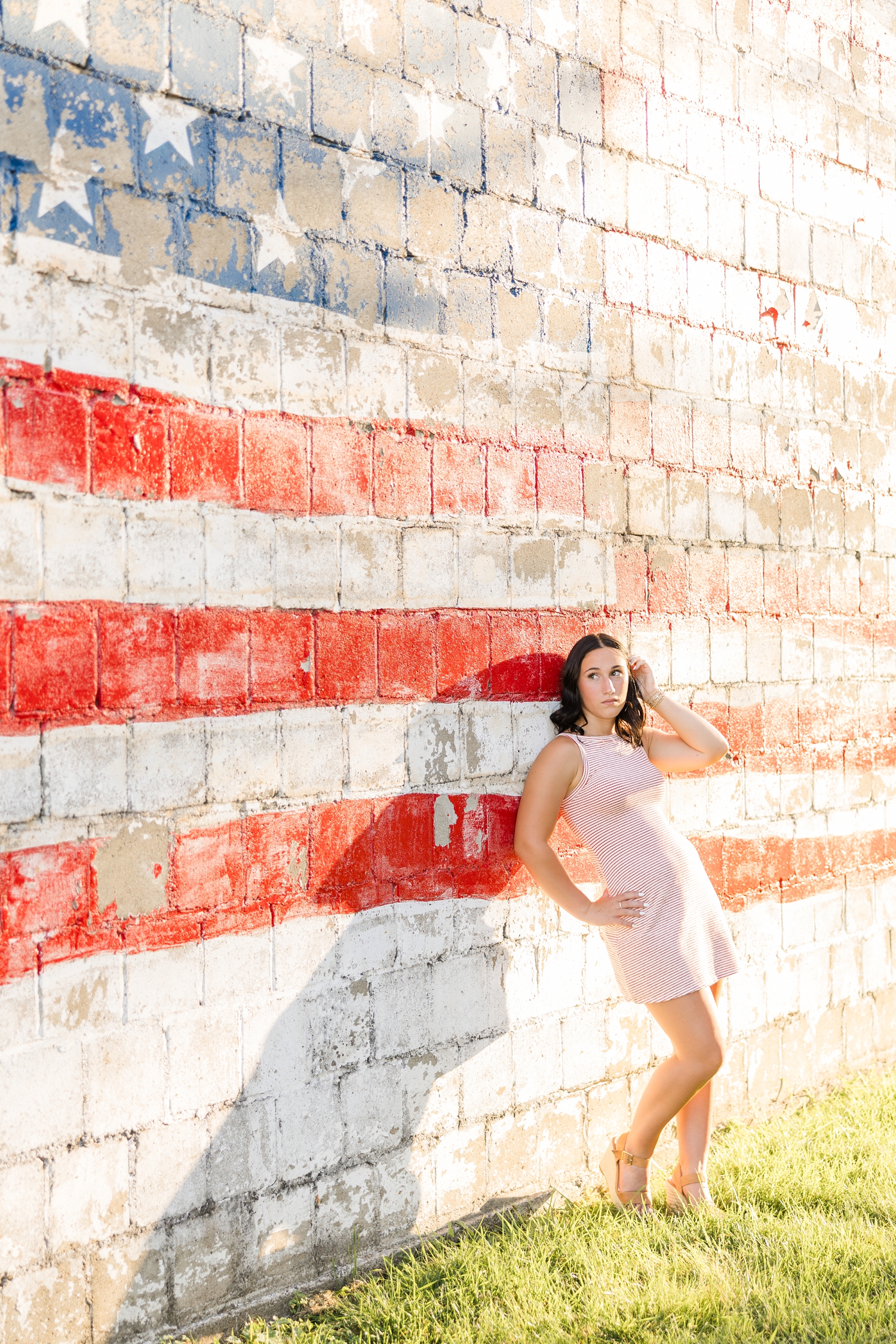 Ivy, wearing a red and white striped dress, leans against a worn white brick building with an American flag painted on it and tucks her hair behind her ears | CB Studio