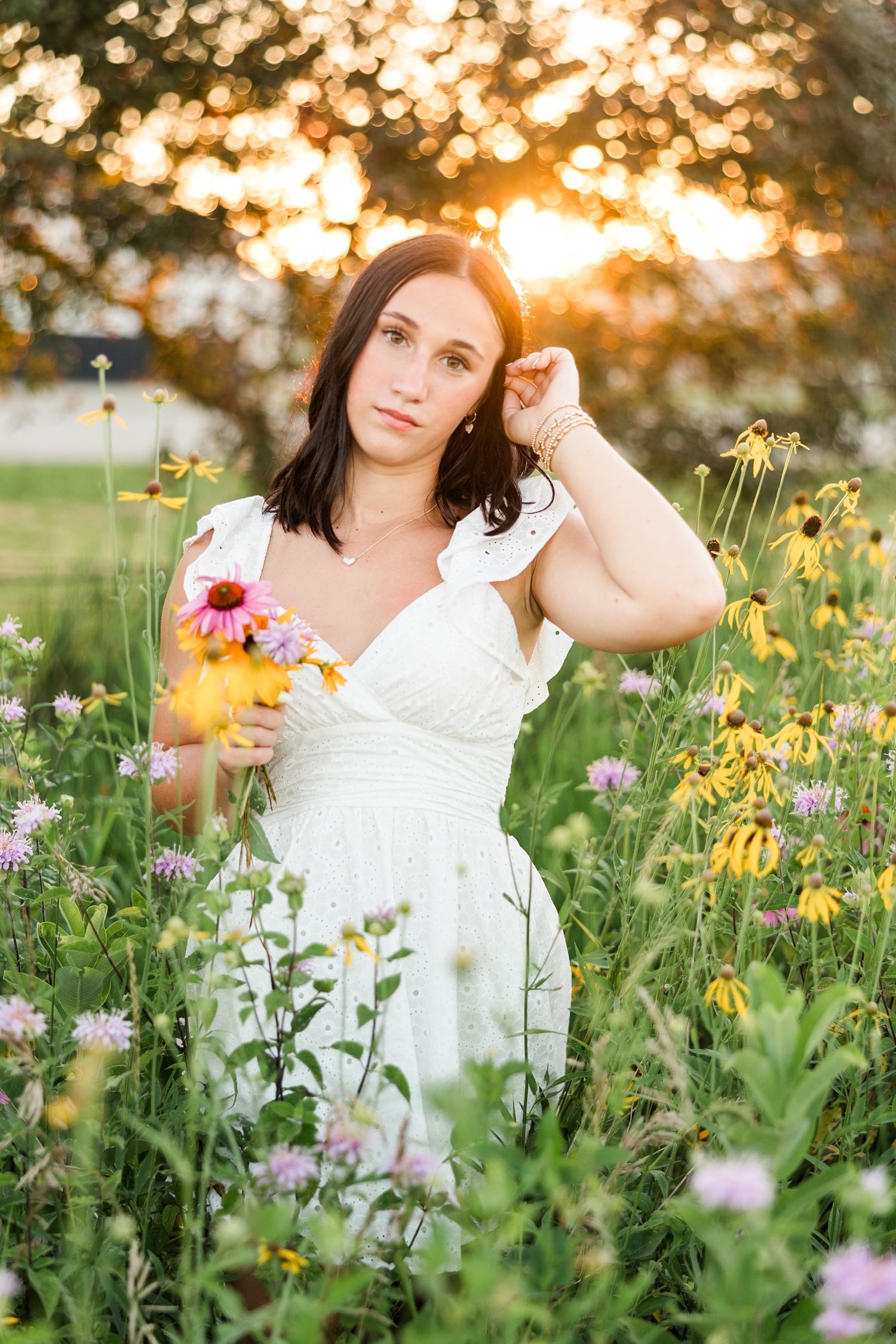 Ivy, wearing a white eyelet dress, stands in a wildflower field at golden hour as she holds a small bouquet and tucks her hair behind her ears | CB Studio
