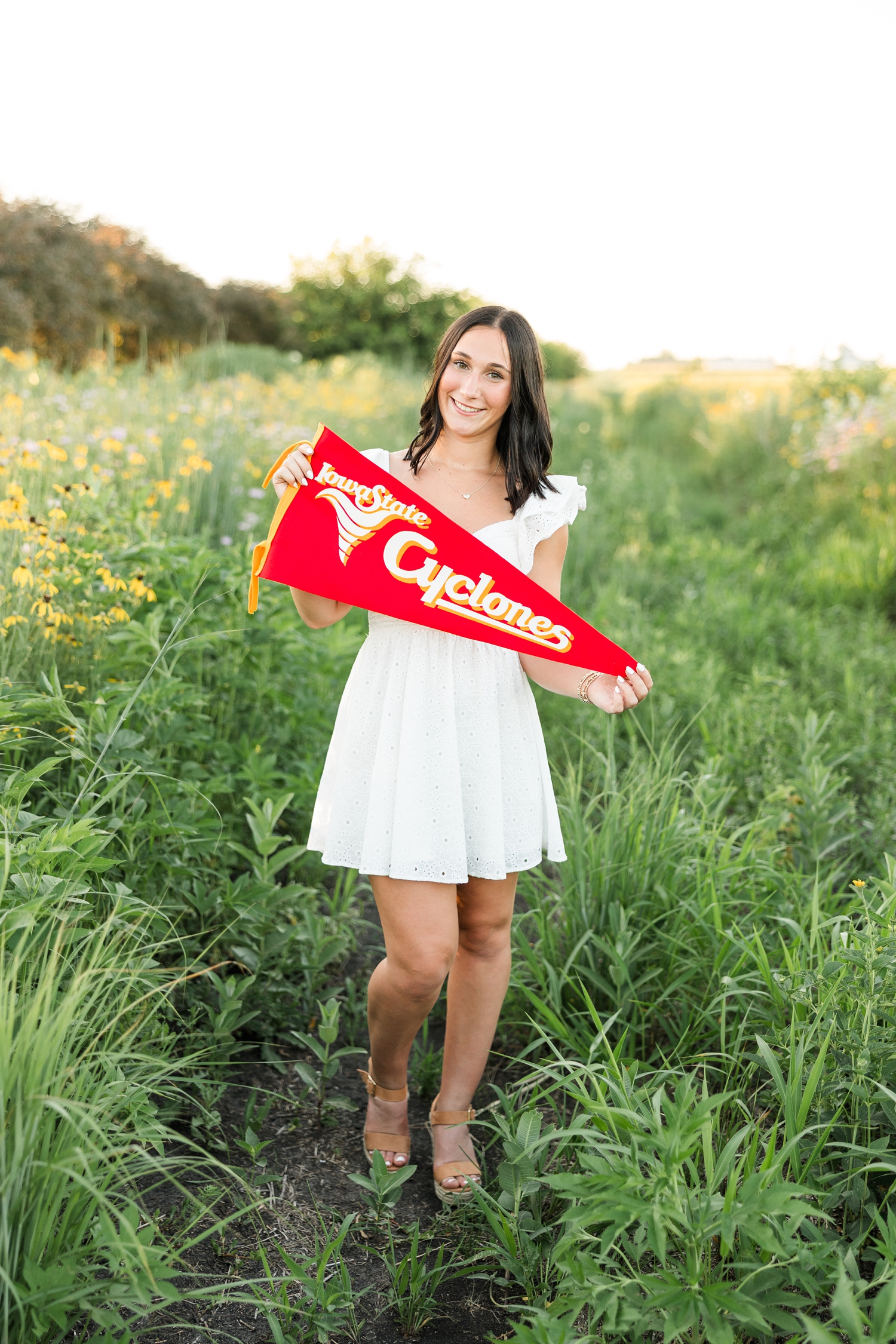 Ivy, wearing a white eyelet dress, stands in a wildflower field at golden hour as she holds a vintage Iowa State Cyclones pennant that belongs to her dad | CB Studio