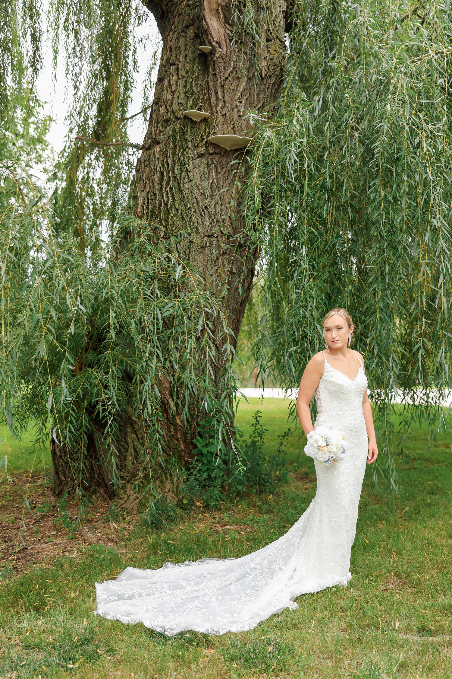 Underneath a willow tree, Alyssa stands hold her wedding bouquet as her wedding gown train trails to the left side | CB Studio