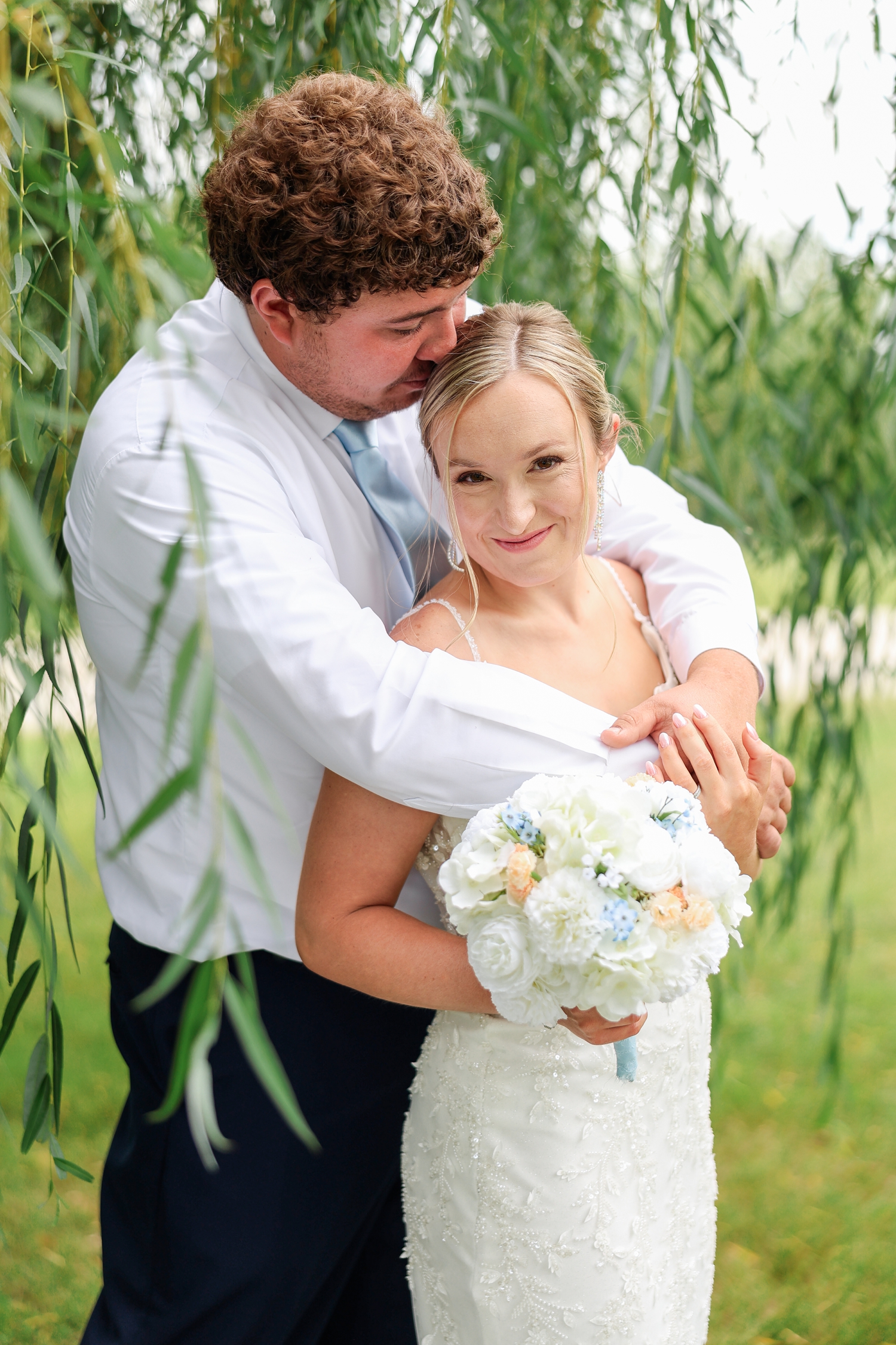 Surrounded by willow branches, Brandon hugs Alyssa from behind and nuzzles the top of her head while she smiles | CB Studio