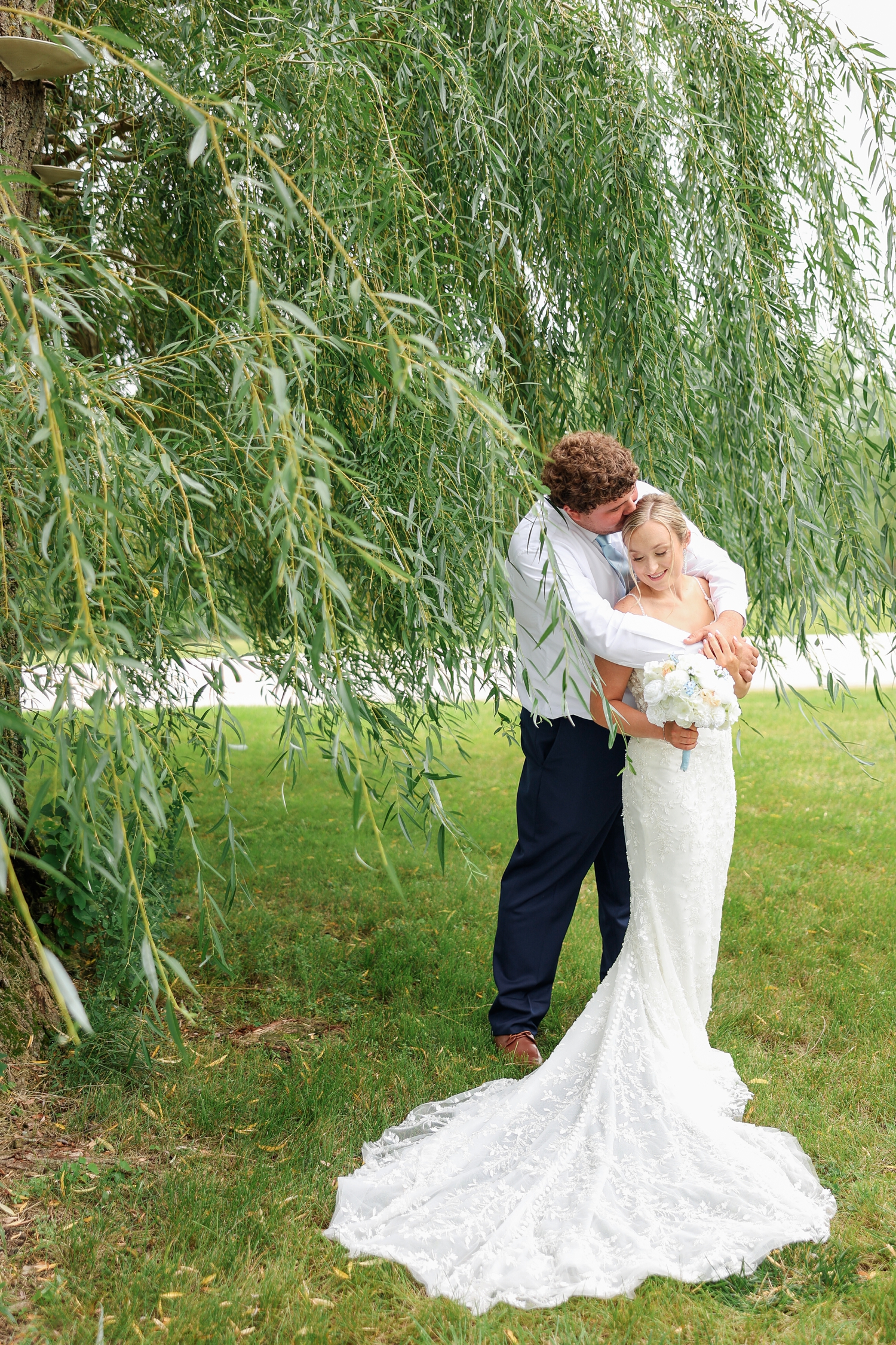 Surrounded by willow branches, Brandon hugs Alyssa from behind and kisses the top of her head while she looks down and smiles | CB Studio