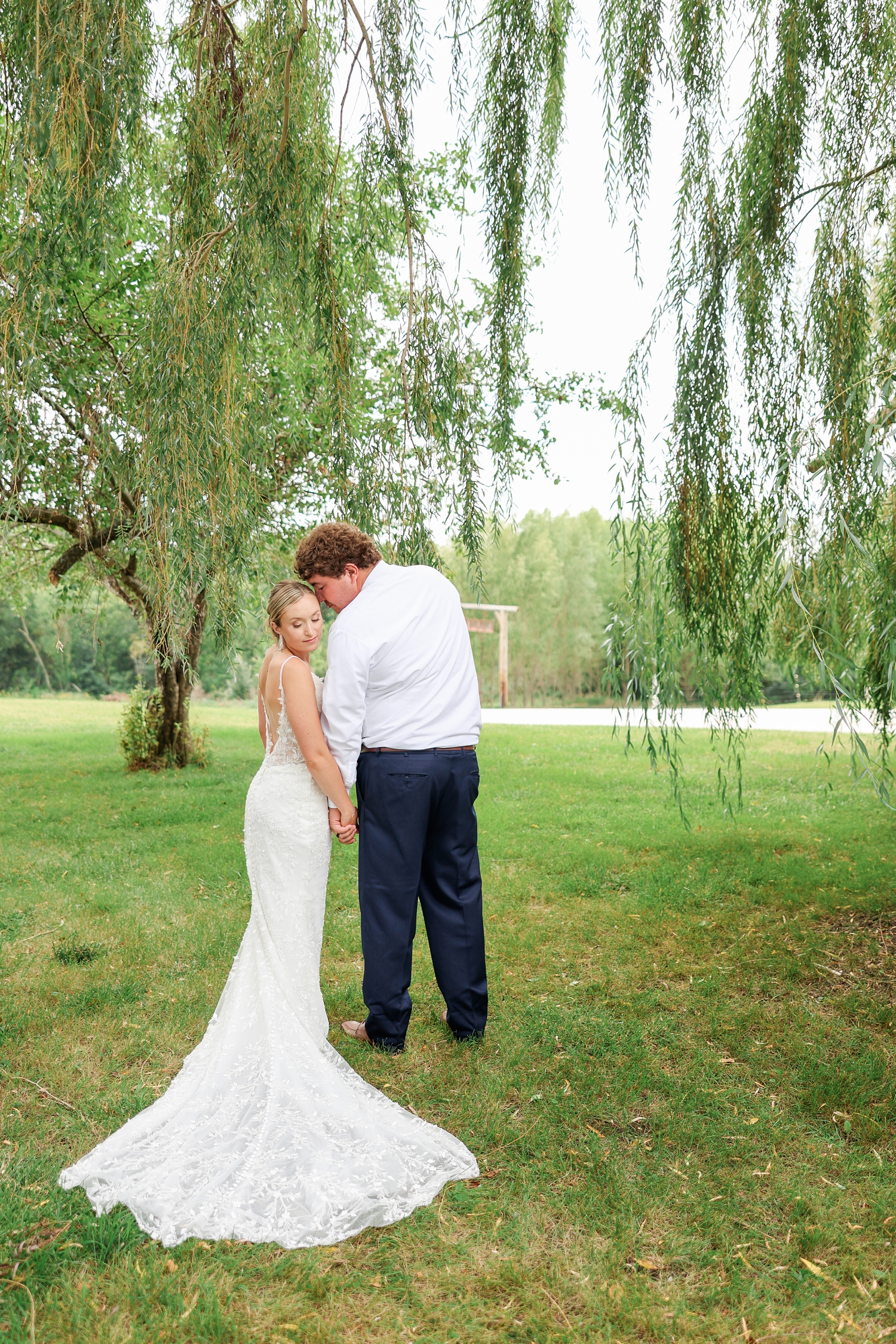 As Alyssa and Brandon walk away holding hands, Alyssa turns back and looks down at her wedding gown train while Brandon nuzzles the side of her face surrounded by draping willow branches | CB Studio