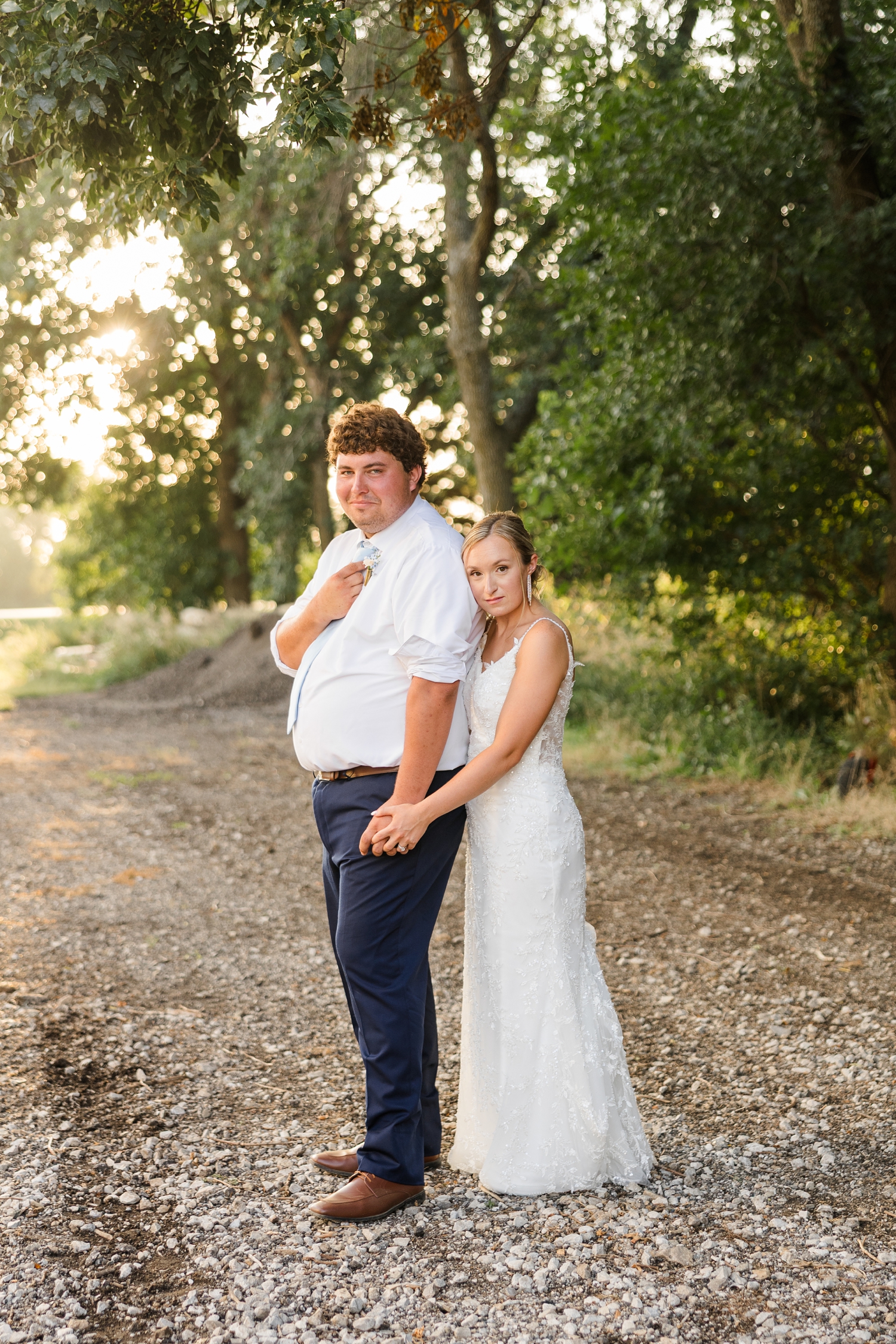 Alyssa stands behind Brandon, holds his hand, and rest her cheek on his shoulder as they both stand on a gravel path along a row of trees at sunset | CB Studio 
