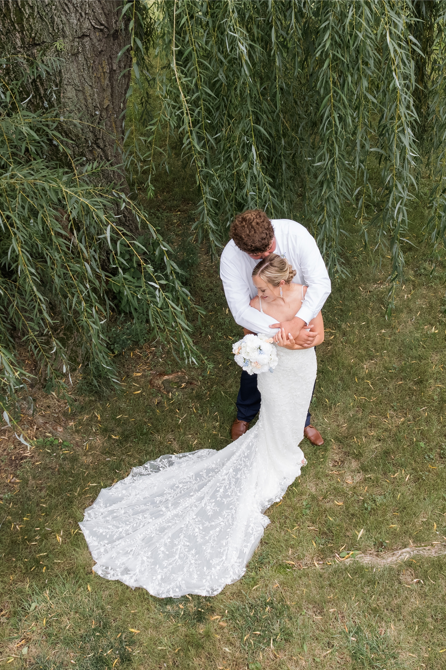 looking from above, Brandon hugs Alyssa from behind and kisses the top of her head while she looks down and smiles all while standing next to a willow tree | CB Studio