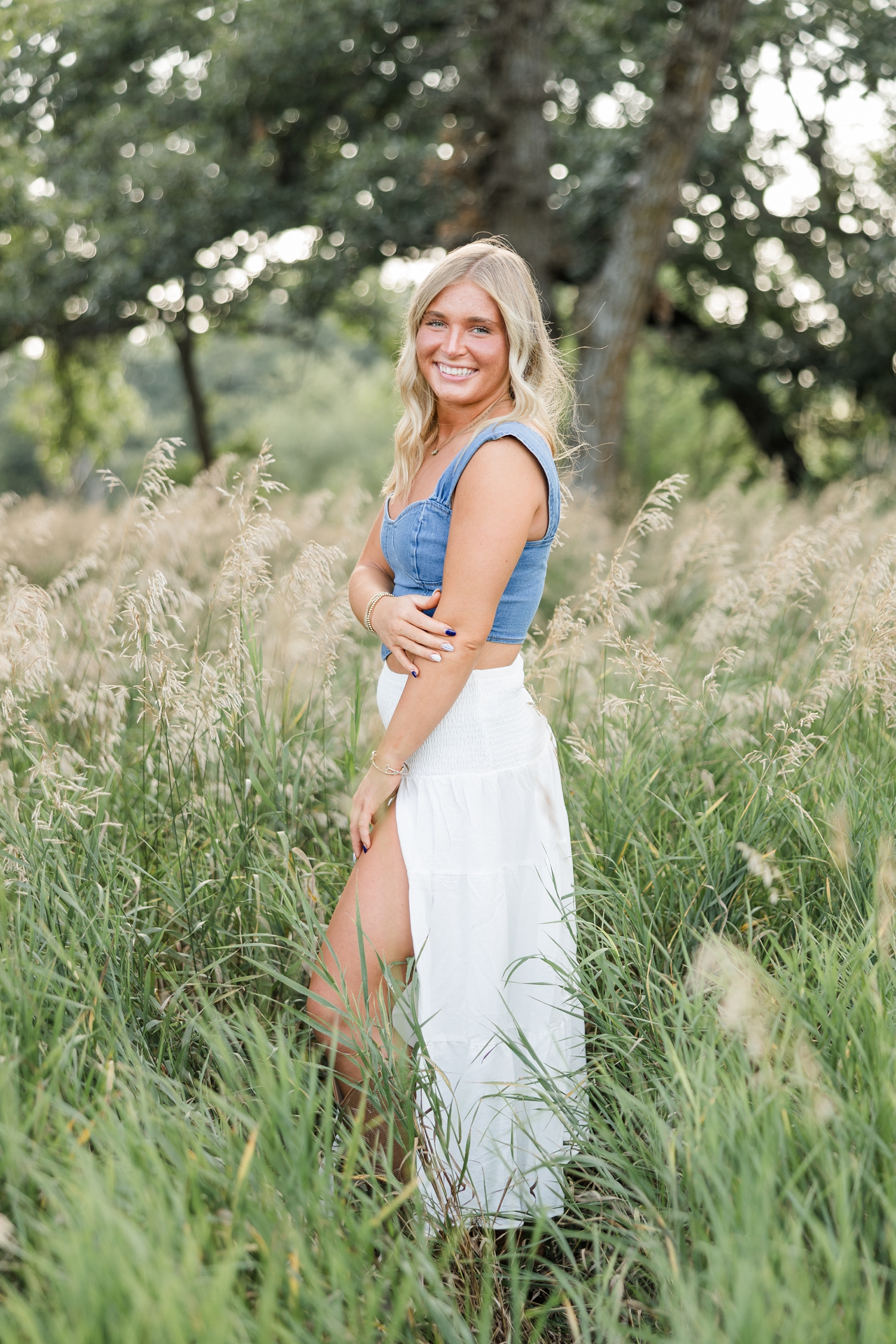 Audrey, wearing a white maxi skirt with a denim tank top, stands a grassy field as she grabs her elbow and smiles as the breeze slowly blows through her hair | CB Studio