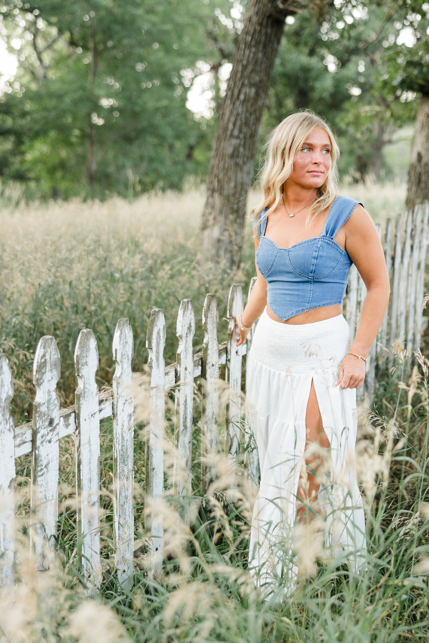 Audrey, wearing a white maxi skirt with a denim tank top, walks through a grassy field filled with oak trees as slowly drags her hand across an old white picket fence | CB Studio