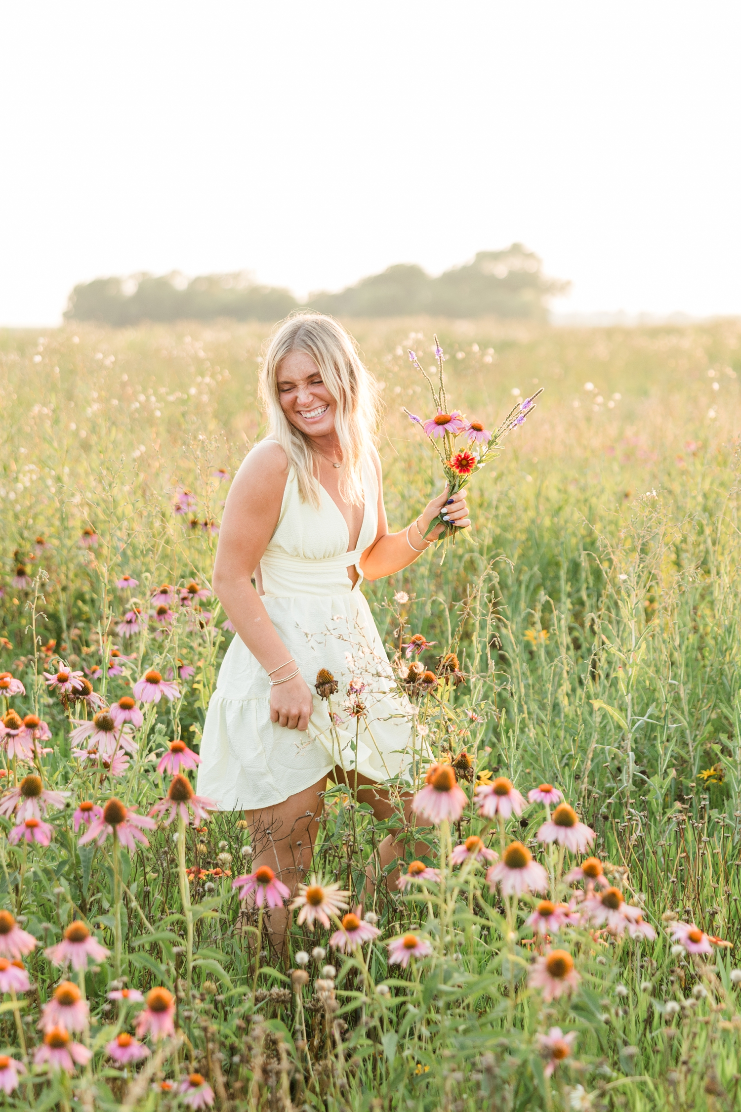 Audrey, wearing a mini butter yellow sundress, walks in a wildflower field during golden hour holding a small flower bouquet as she giggles and looks down | CB Studio