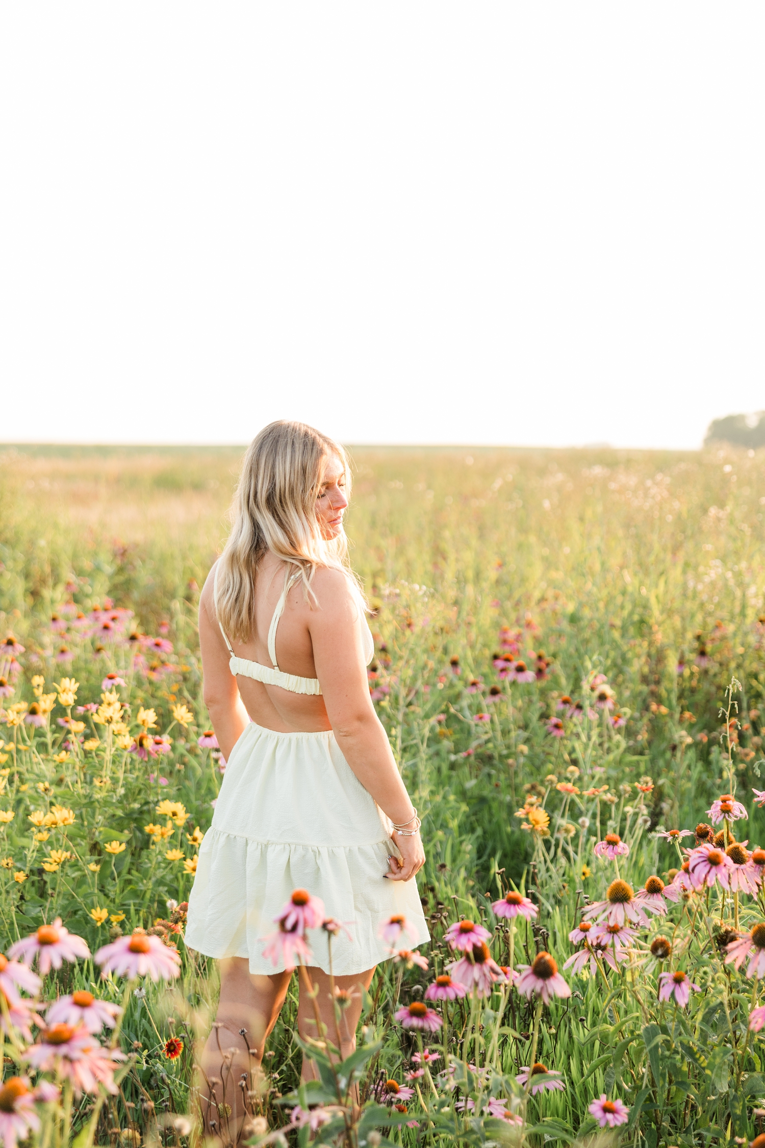 Audrey, wearing a mini butter yellow sundress, stands in a wildflower field during golden hour facing away from the camera and looking back and down over her shoulder | CB Studio