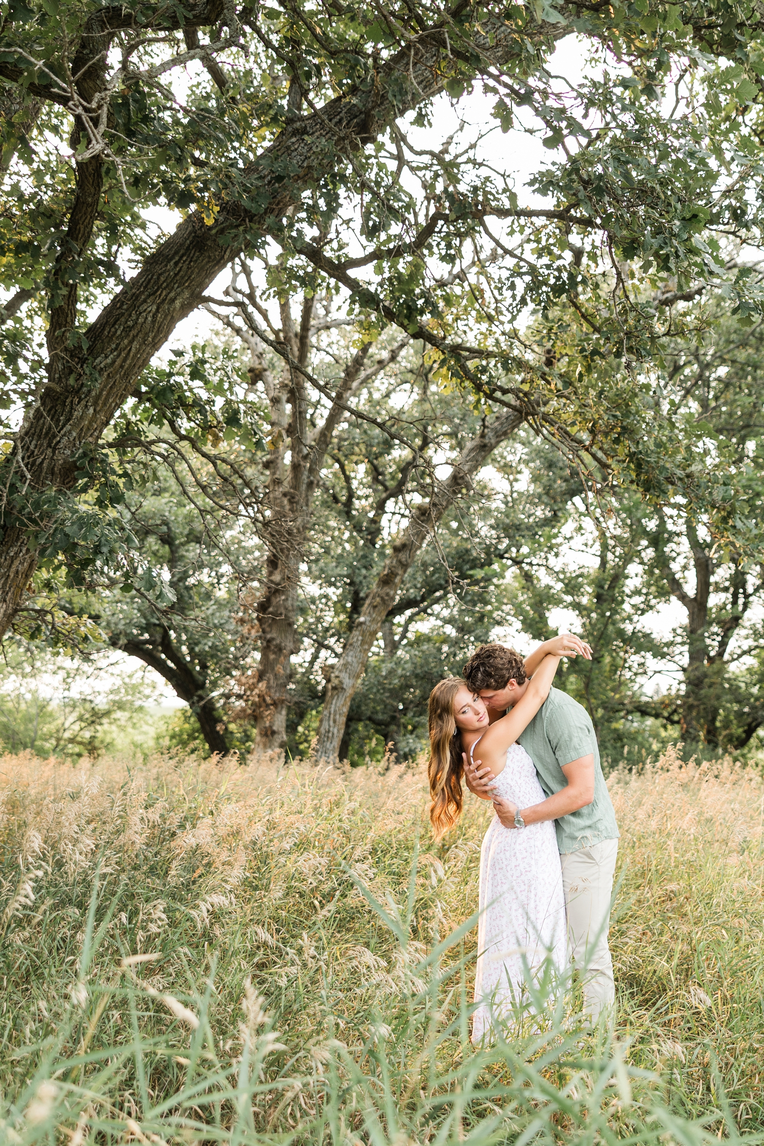 Tyler dips Deni back as he nuzzles her cheek and her arms are crossed straight over his shoulders in the middle of a grass pasture as oak trees frame around them | CB Studio