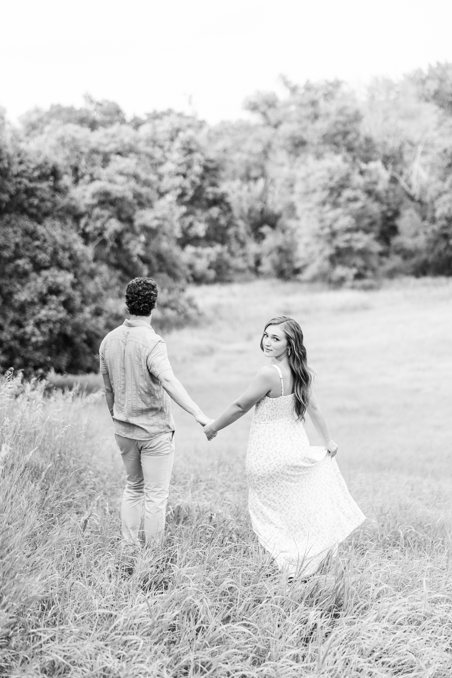 Deni and Tyler walk hand and hand away from the camera down a hill in a grass pasture as Deni looks back at the camera | CB Studio