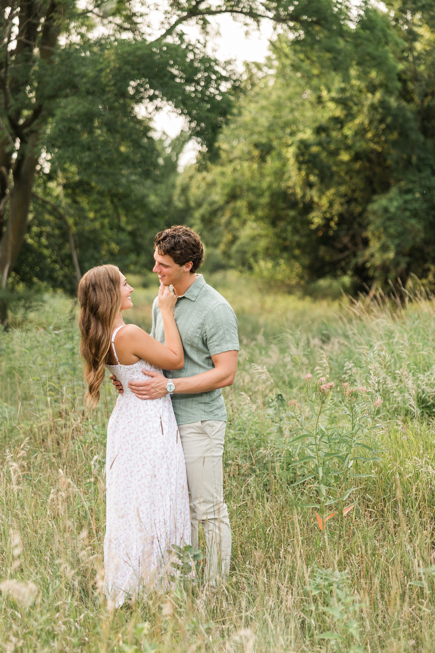 Deni gently holds Tyler's chin as they smile at each other in the middle of a grass pasture lined with trees in the background as butterflies dance in the foreground | CB Studio