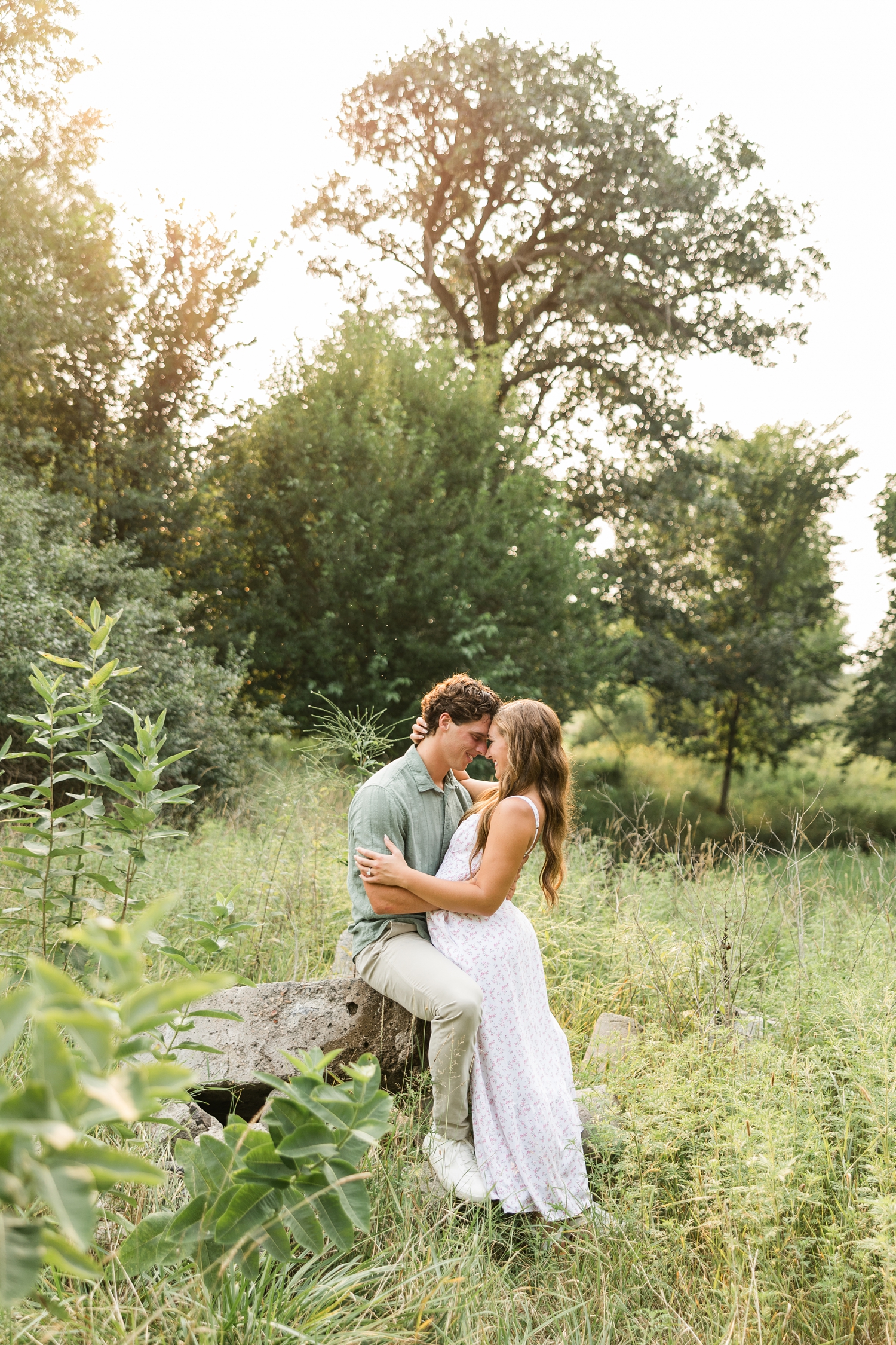 Tyler, wearing a green button down and khaki pants, sits on a rock formation in a grass pasture as Deni, wearing a white dress with a pink ditsy floral pattern, embraces him and they rest their foreheads together | CB Studio