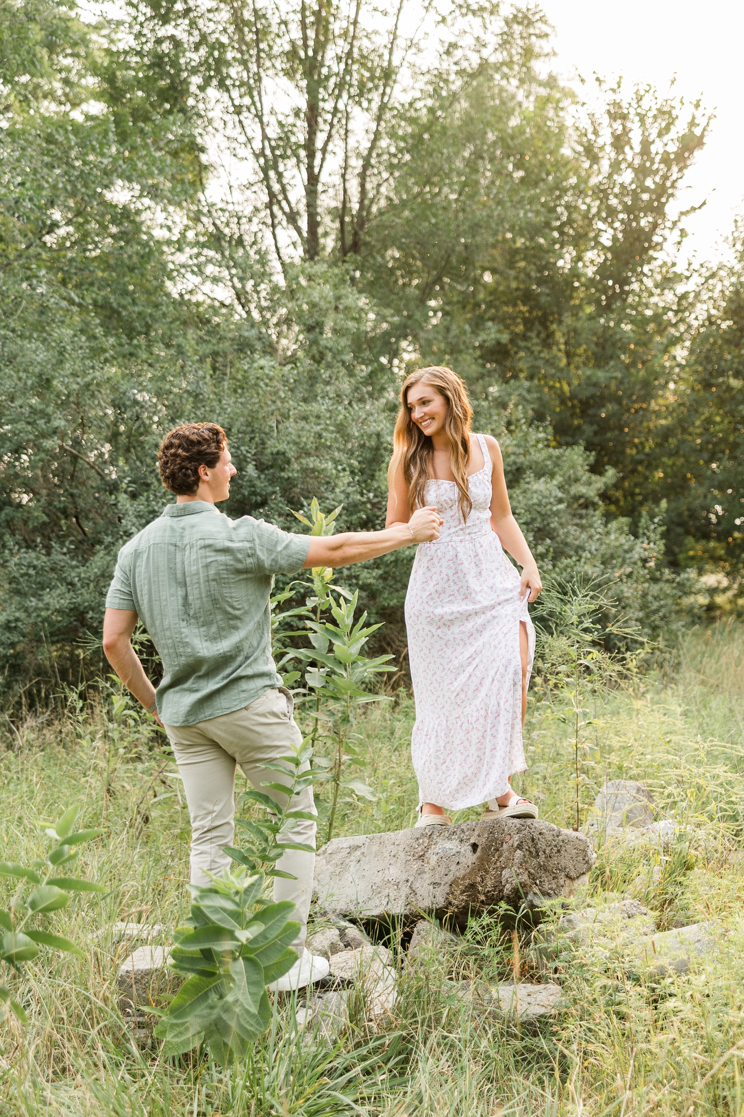 Deni, wearing a white dress with a pink ditsy floral pattern, walks on top of some rocks in a grass pasture while Tyler, holds her hand and helps her across the rock as they smile at each other | CB Studio