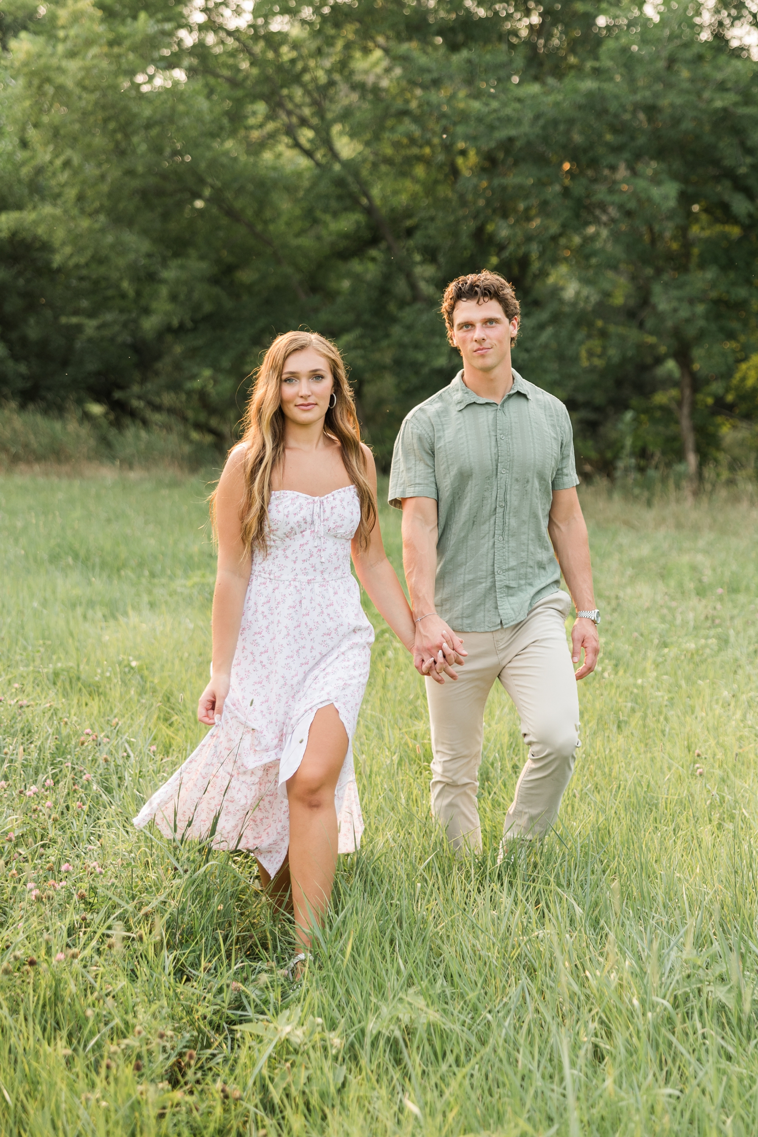 Deni, wearing a white dress with a pink ditsy floral pattern, walks hand in hand with Tyler, wearing a green button down and khaki pants, in the middle of a grass pasture with trees lining the background | CB Studio