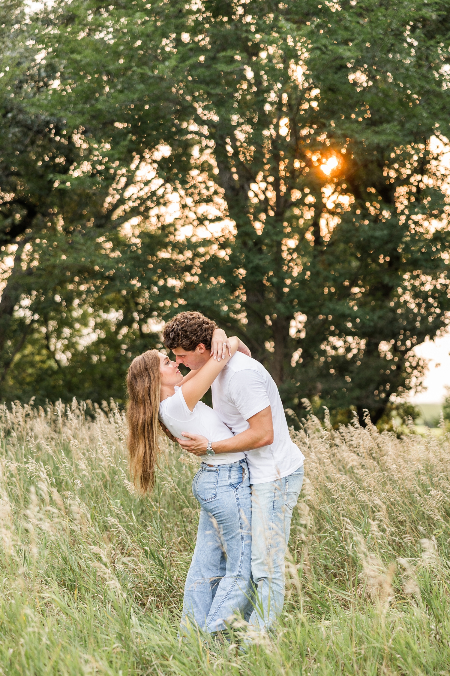 Tyler dips Deni (both wearing jeans and a white t-shirt) back as they touch noses in the middle of a grass pasture with the sun setting behind the oak trees in the background | CB Studio