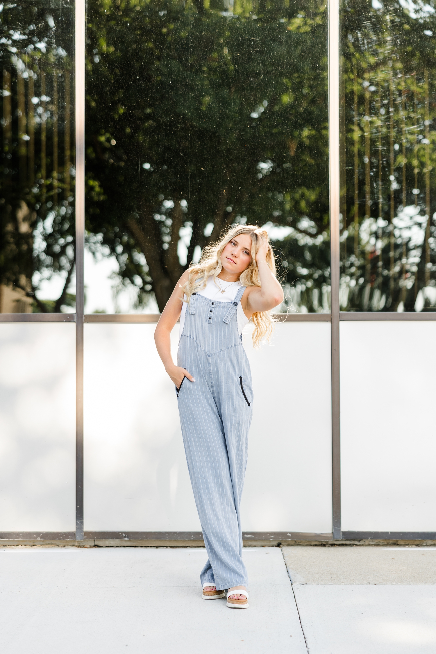 Emma, wearing a mock denim jumpsuit and white tank, stands in front of a mirrored building in downtown Algona as she has one hand in her pocket and the other in her hair | CB Studio