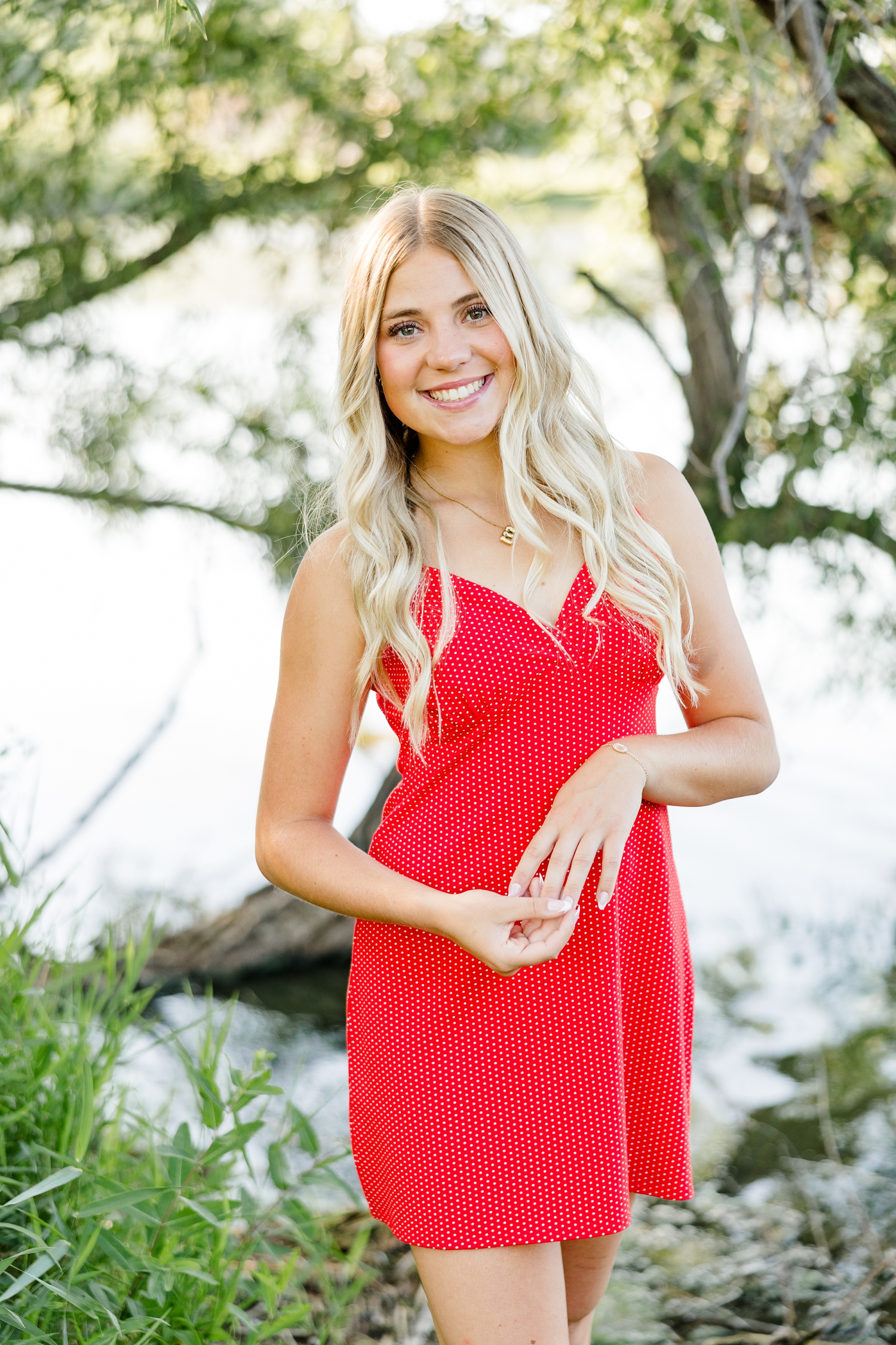 Emma, wearing a red dress with small white polka dots, stands gently holding her hands in front of Smith Lake with trees draping in the background | CB Studio