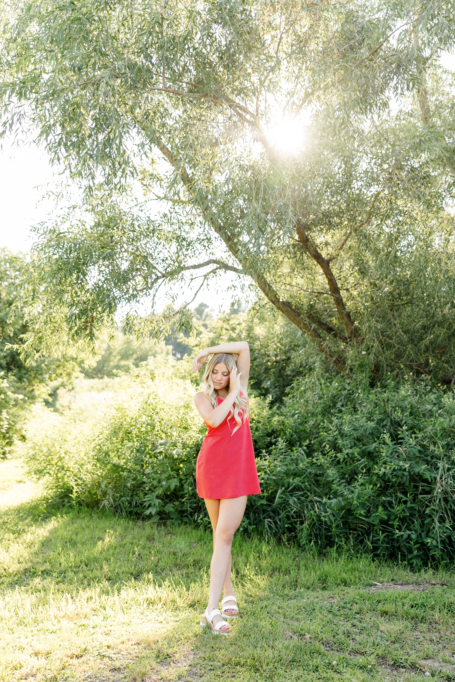 Emma, wearing a red dress with small white polka dots, stands in a grass pathway with her hands resting on top of her head as the sun shines through the trees in the background | CB Studio