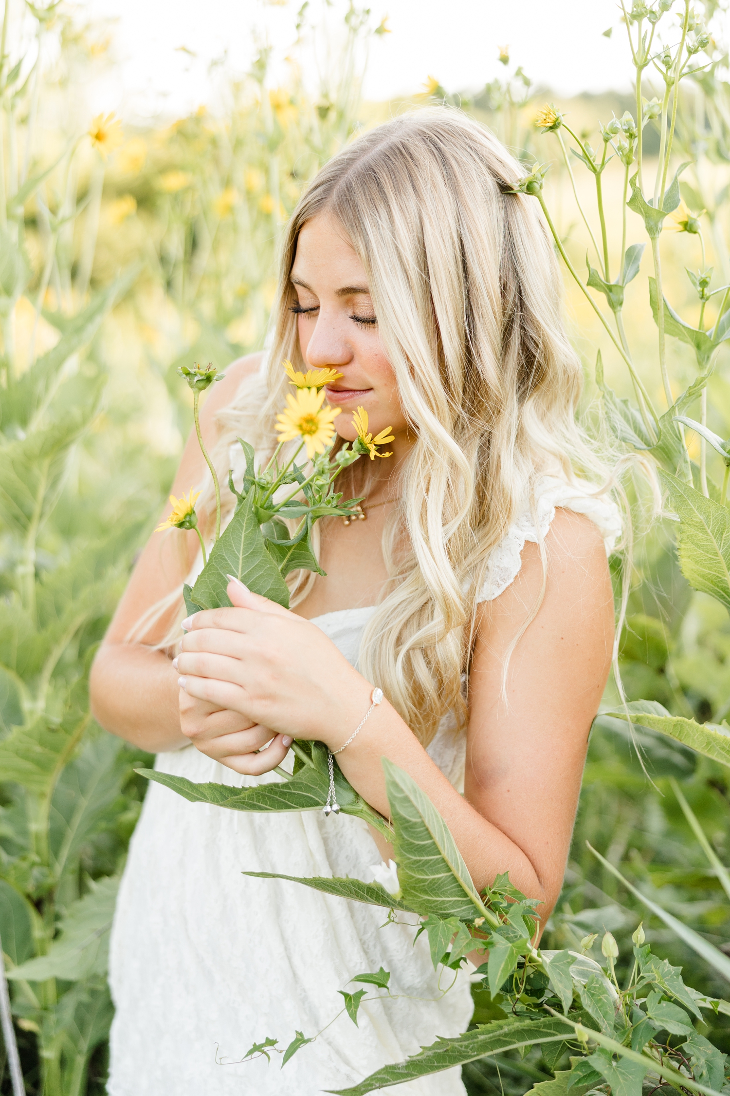 Emma, wearing a cream lace dress, stands surrounded in a yellow cup flower field while she brings a flower to her nose to smell it | CB Studio