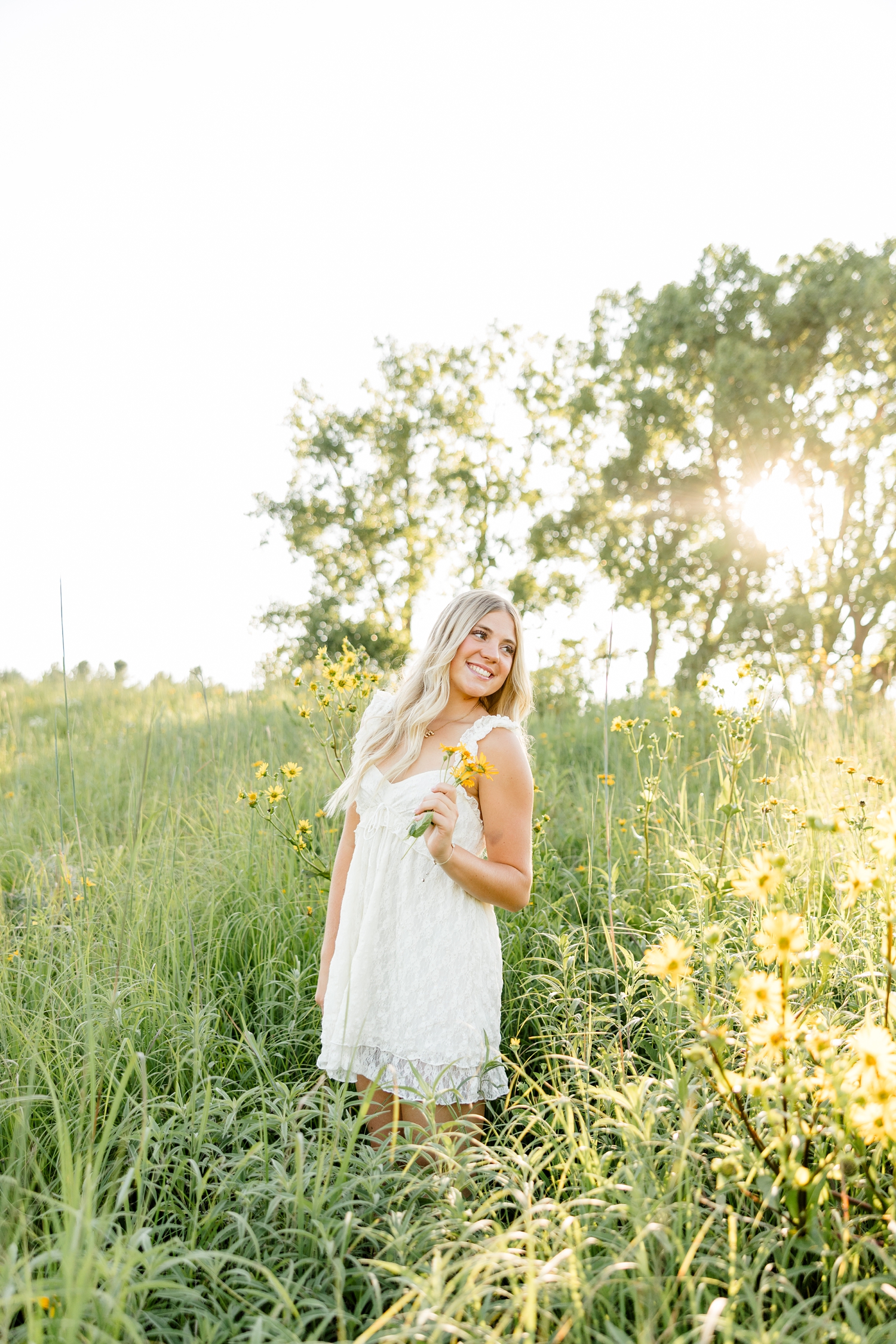 Emma, wearing a cream lace dress, stands in a yellow flower field at golden hour and looks over her shoulder to the right while holding a small bouquet of flowers | CB Studio