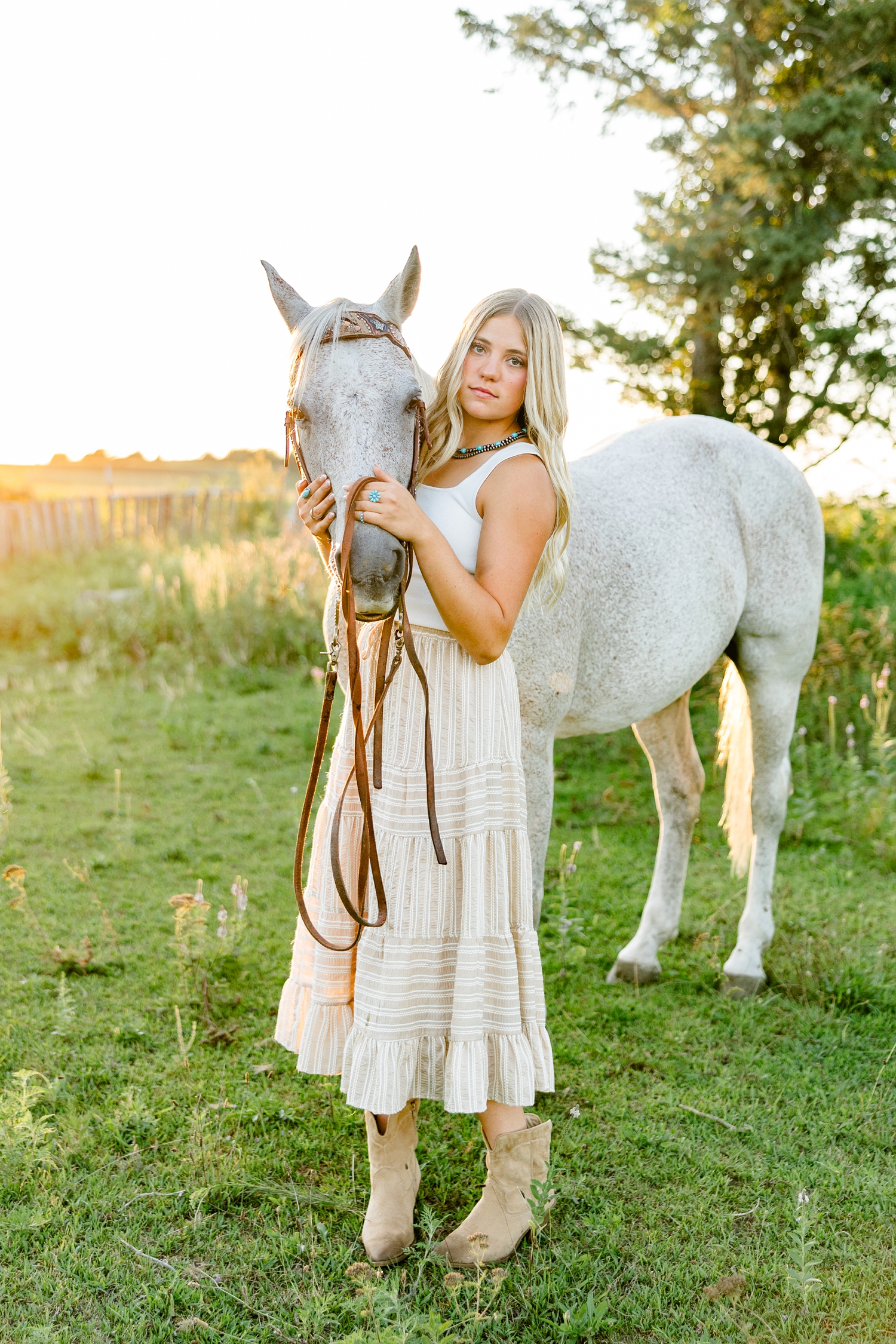 Emma, wearing a cream tank top and tan striped skirt, stands holding the nose of her horse in a pasture during golden hour | CB Studio