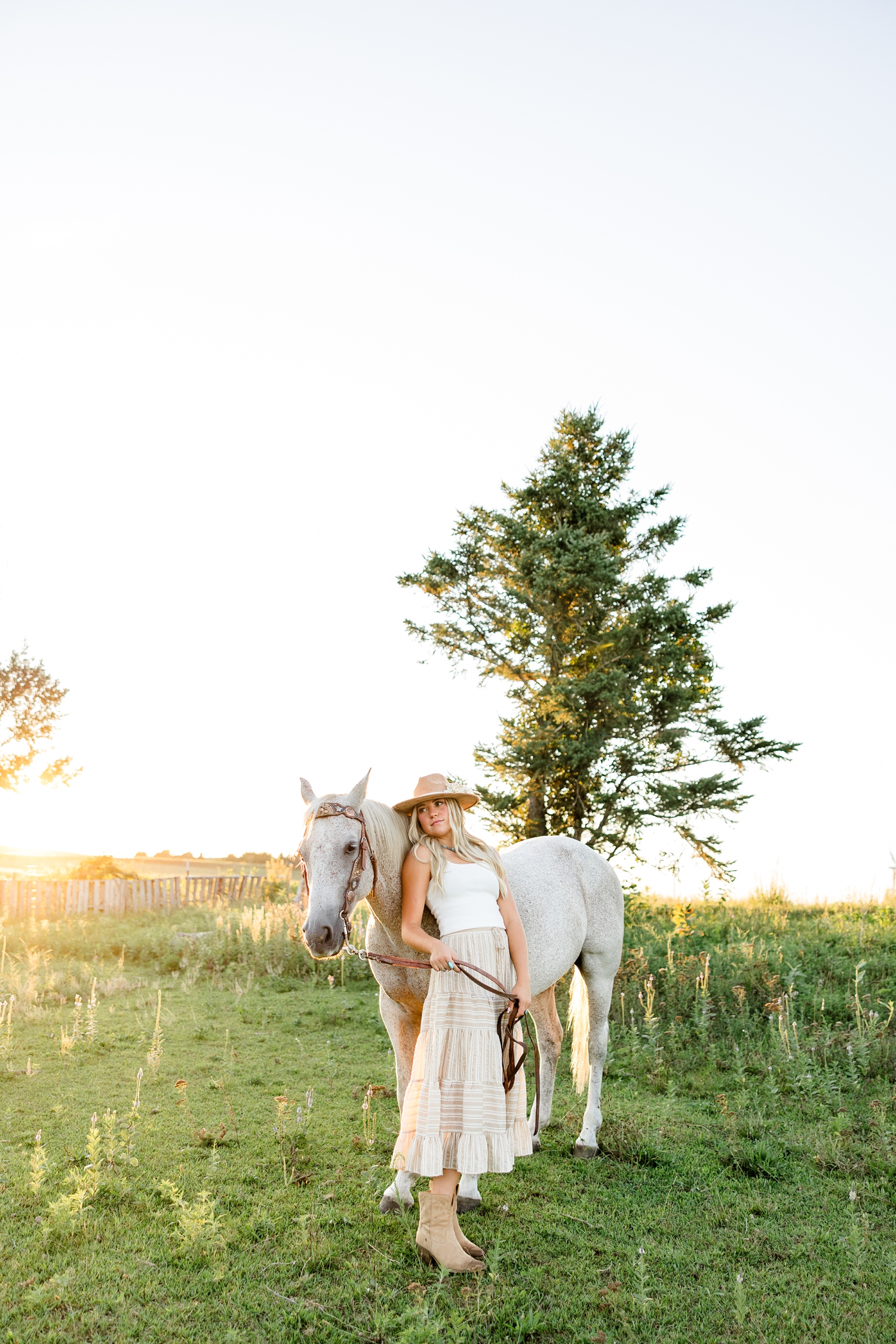 Emma, wearing a cream tank top, tan striped skirt and tan cowgirl hat, leans her back against the shoulder of her tan horse in a pasture during golden hour | CB Studio